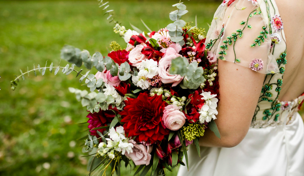 A stunning bridal bouquet featuring deep red and blush pink roses with white flowers and eucalyptus against a green lawn.