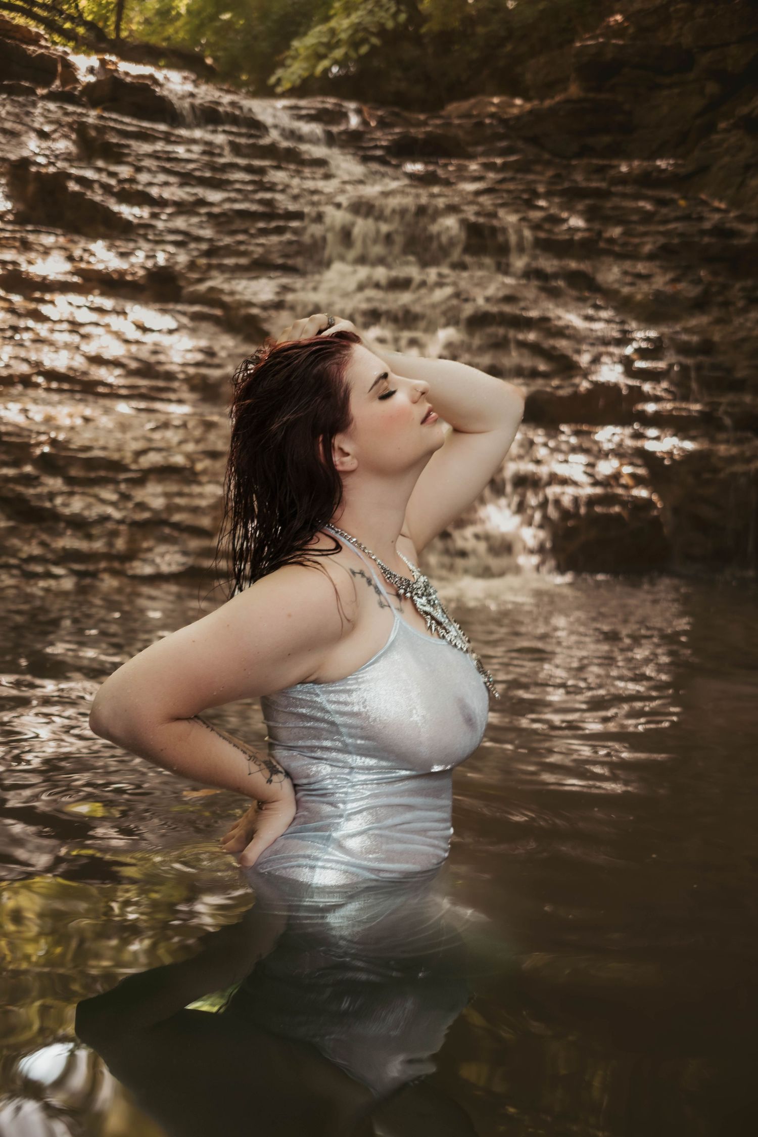 Ethereal portrait in silver dress standing in waterfall pool with dramatic lighting.
