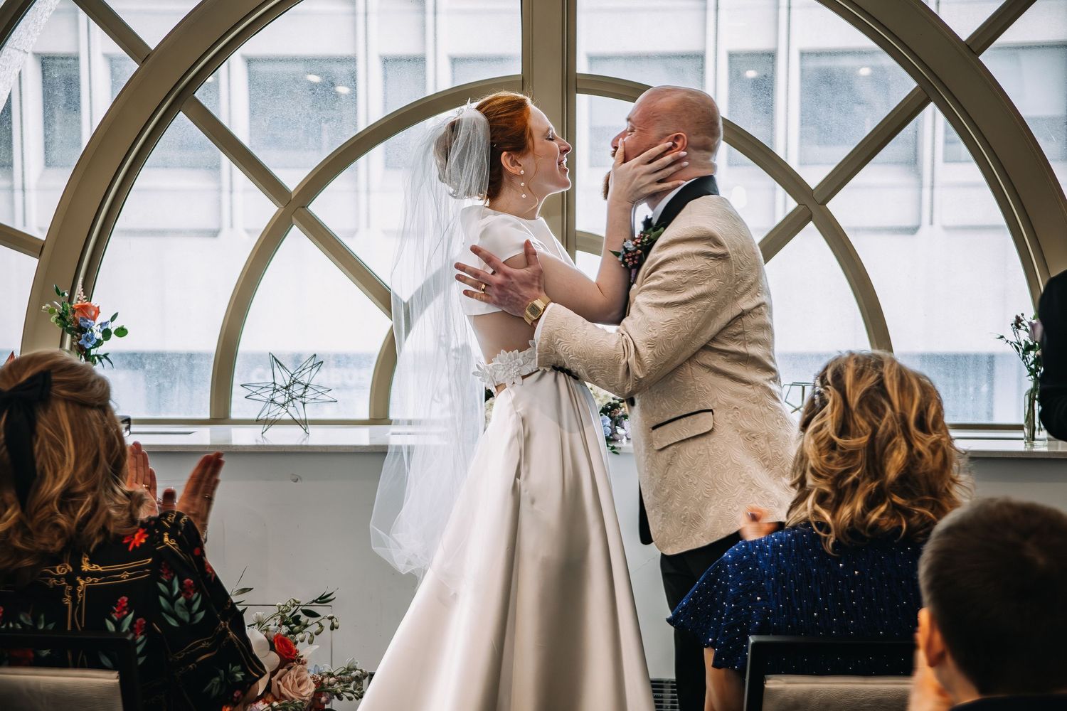 Bride and groom share first kiss at wedding ceremony before arched windows, guests seated in foreground.