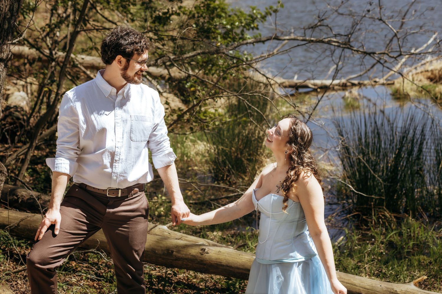 Couple holding hands and smiling at each other near a wooden fence with a lake and trees in the background.