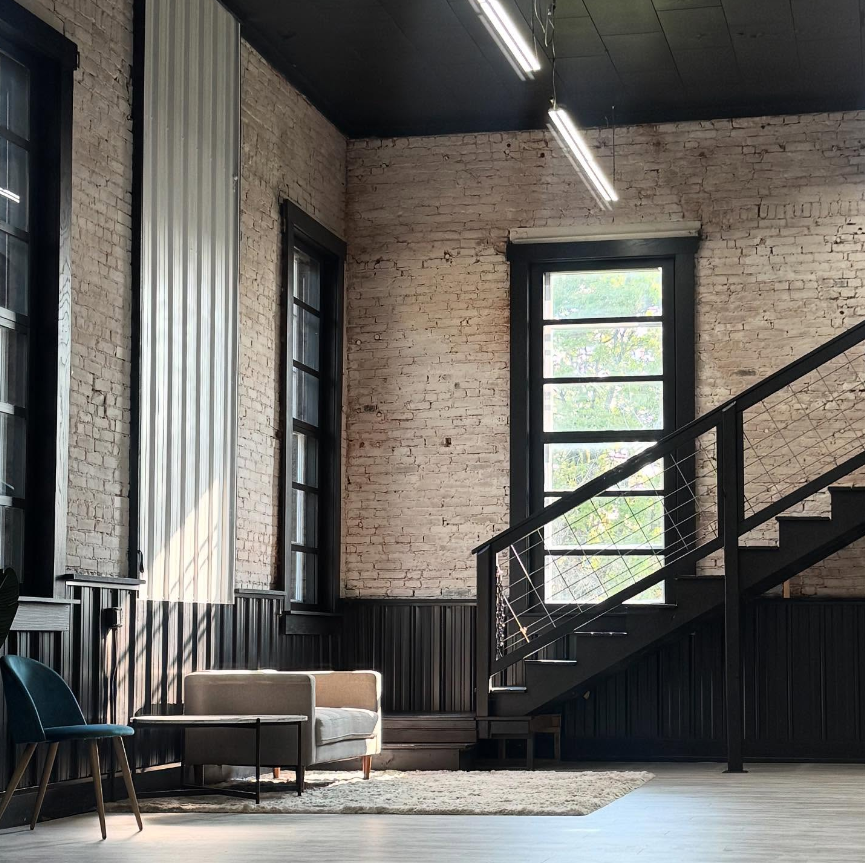 Modern industrial loft space with exposed brick walls, large windows, and sleek black accents in an open floor plan design.