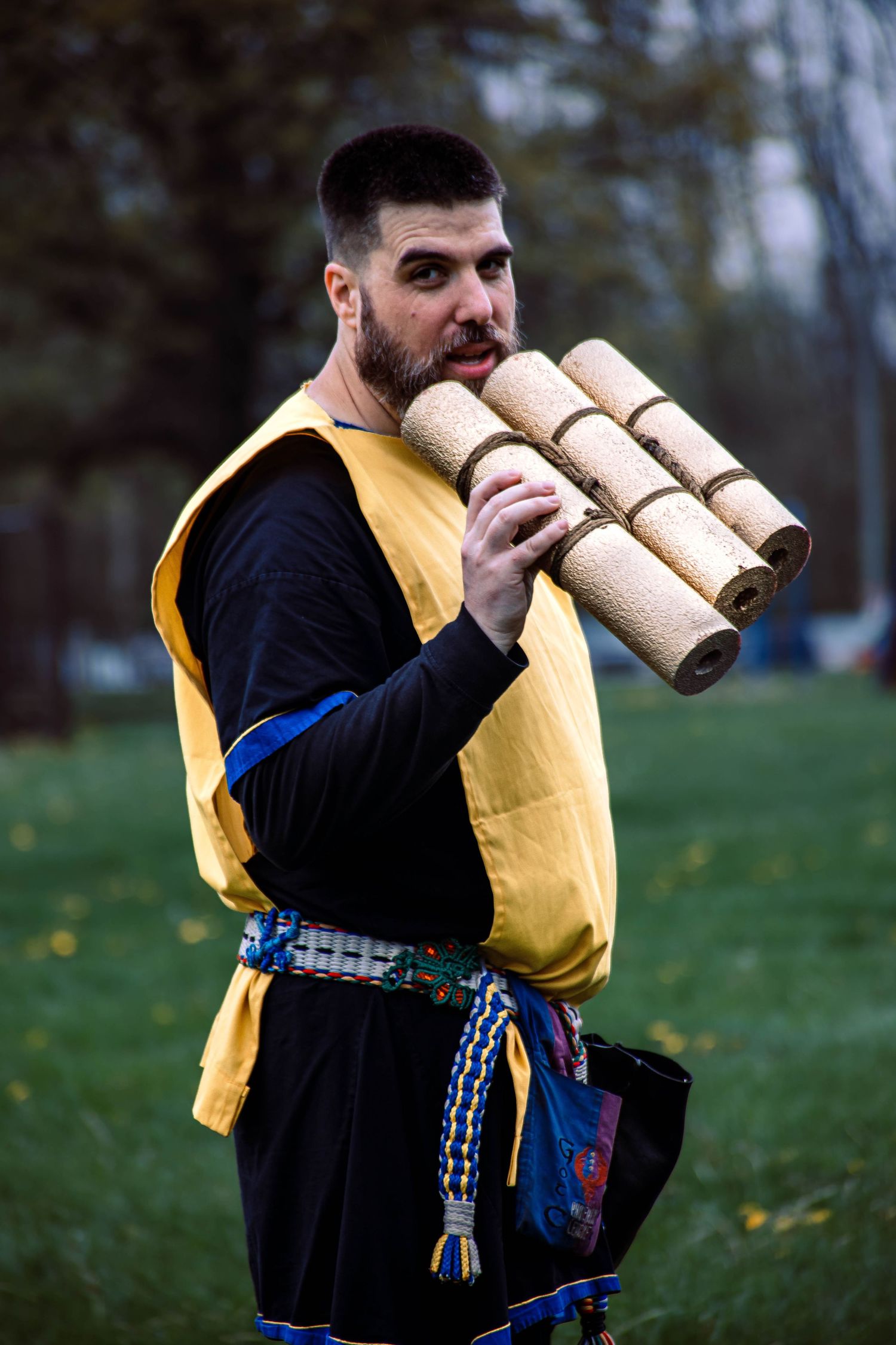 Person wearing black and yellow varsity jacket playing traditional wooden wind instrument in grassy outdoor setting.