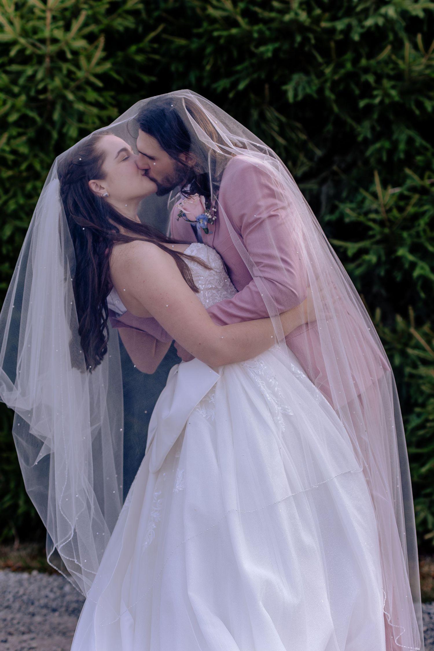 A couple in wedding attire share a romantic kiss outdoors, with the bride in white and groom in pink.
