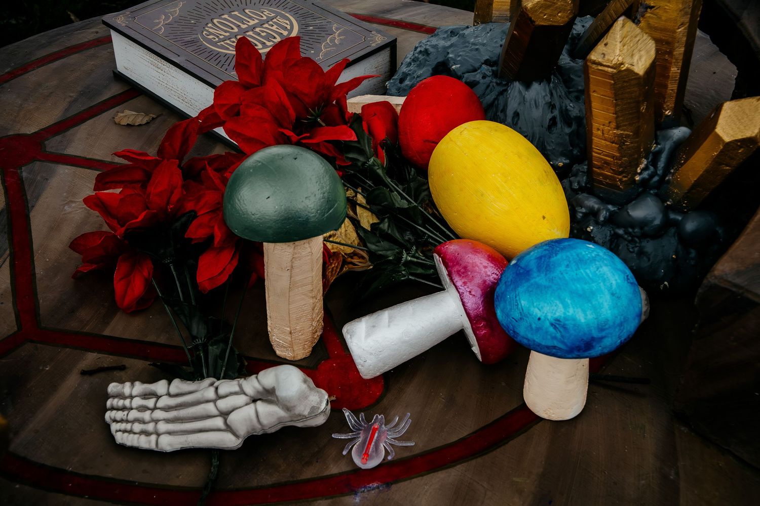Colorful decorative mushrooms, red flowers, and a skeleton hand on a table with occult decor.