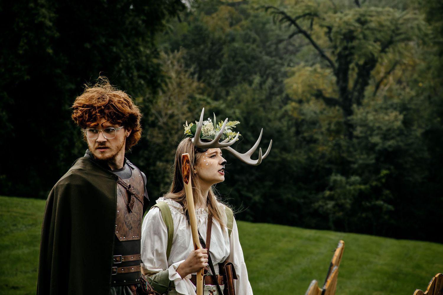 Two costumed performers in medieval fantasy attire, one wearing antler crown with flowers, pose outdoors on green lawn.
