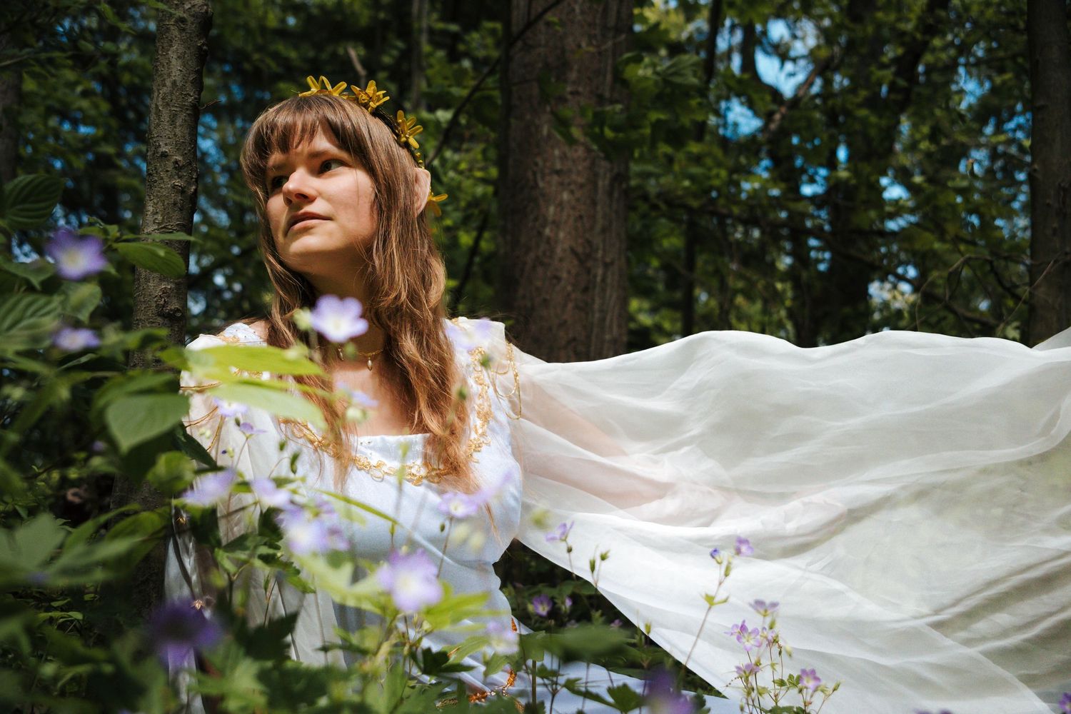 A person in a flowing white dress twirls gracefully in a sunlit forest garden with purple flowers blooming nearby.