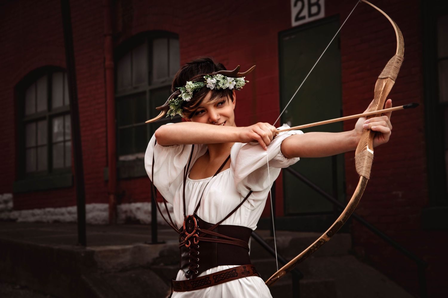 Woman in medieval costume with floral crown drawing a wooden bow and arrow outdoors near a brick building.