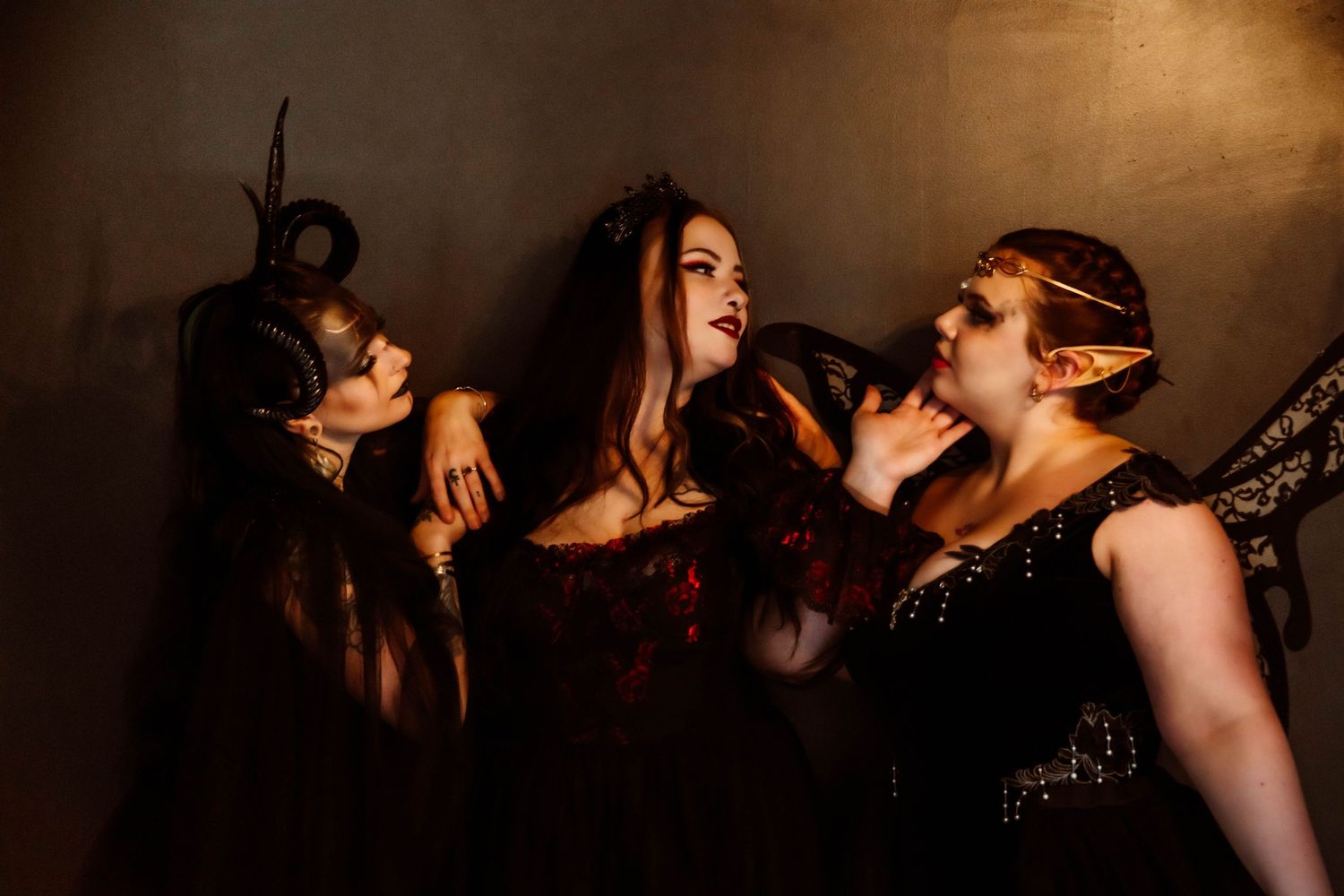 Three women in dark fantasy costumes and masks pose dramatically against a moody golden-brown wall in theatrical Halloween attire.