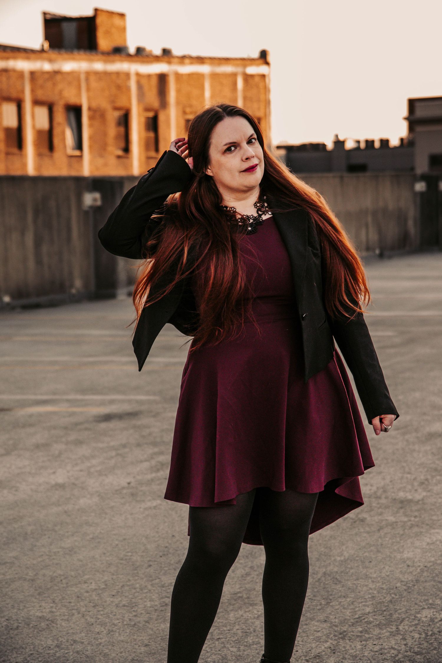 Woman in burgundy dress and black blazer posing on rooftop with long red hair, wearing black tights and statement necklace.