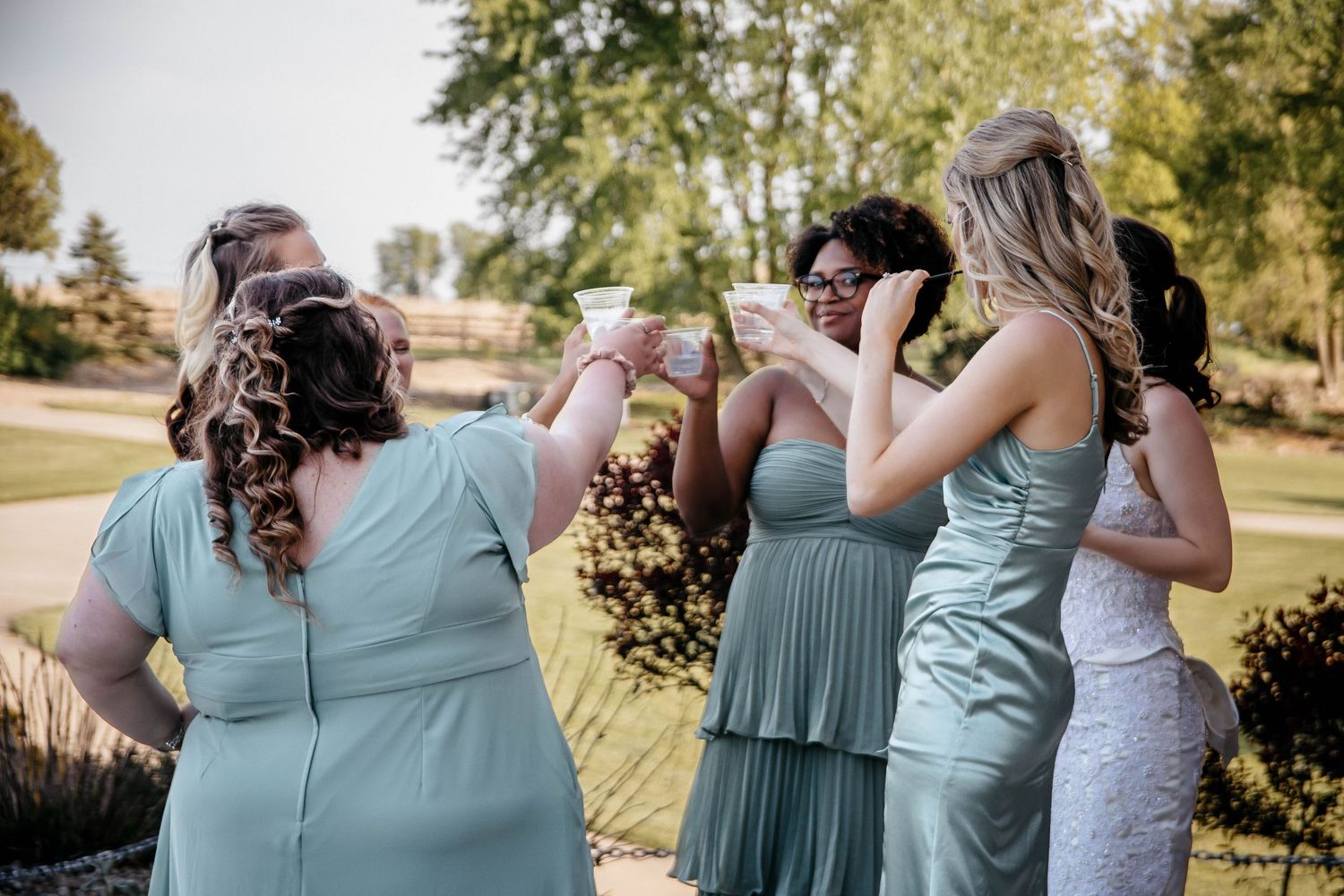Bridesmaids in sage green dresses toasting outdoors at a wedding celebration on a sunny day.