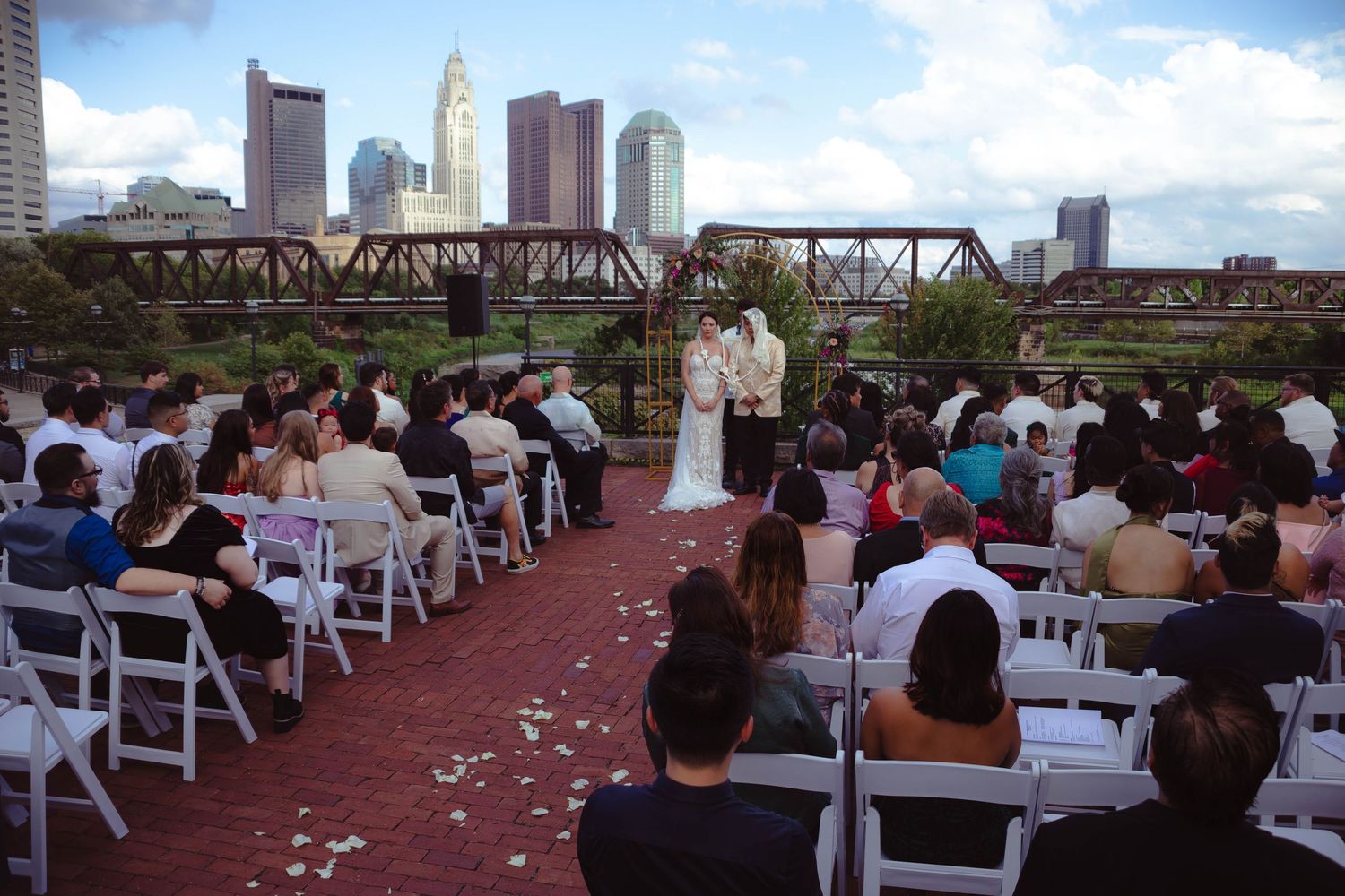 Outdoor wedding ceremony with guests seated in white chairs, bride and groom at altar, city skyline backdrop.