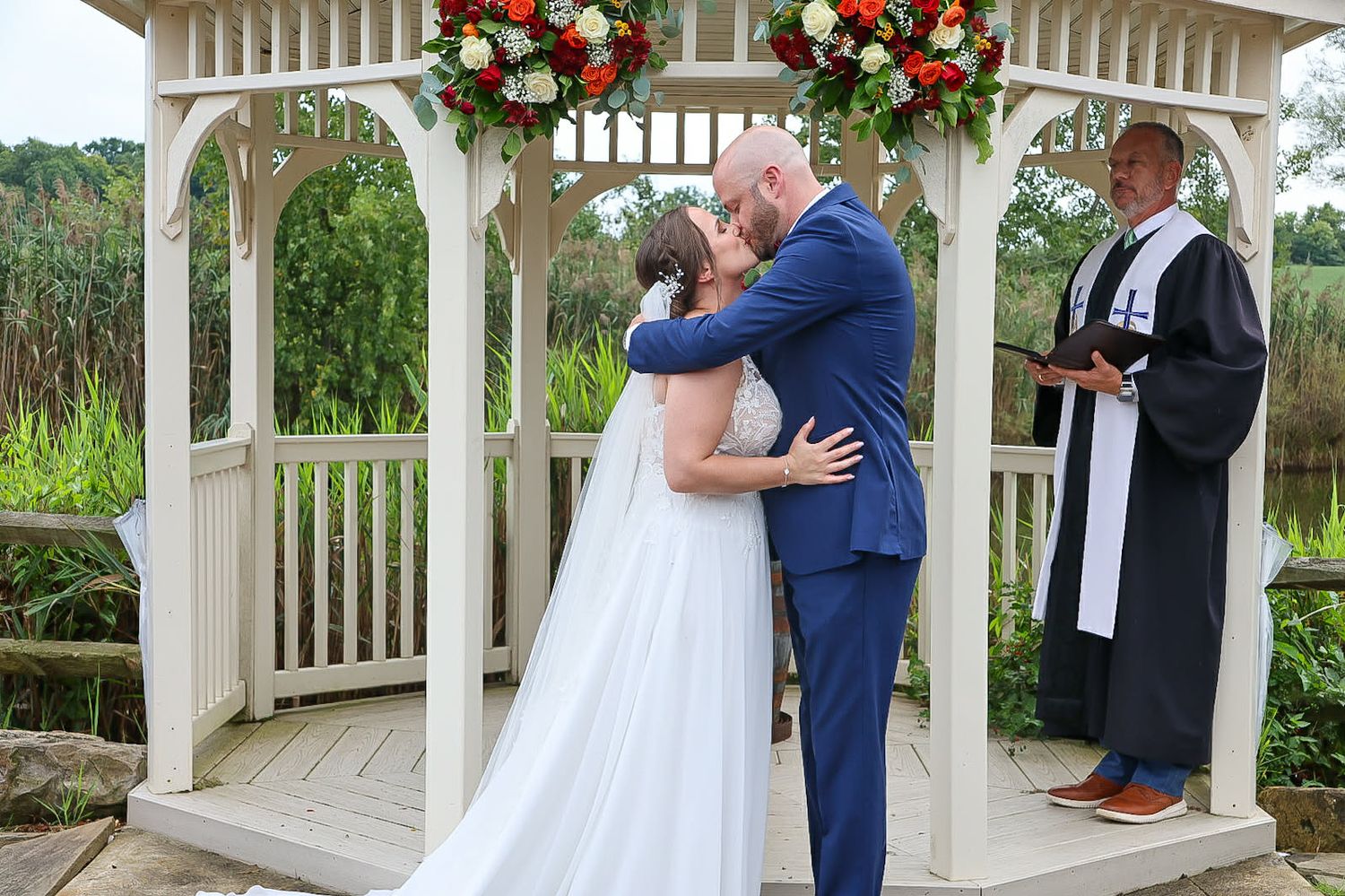 Bride and groom share first kiss under floral gazebo with officiant during outdoor wedding ceremony.