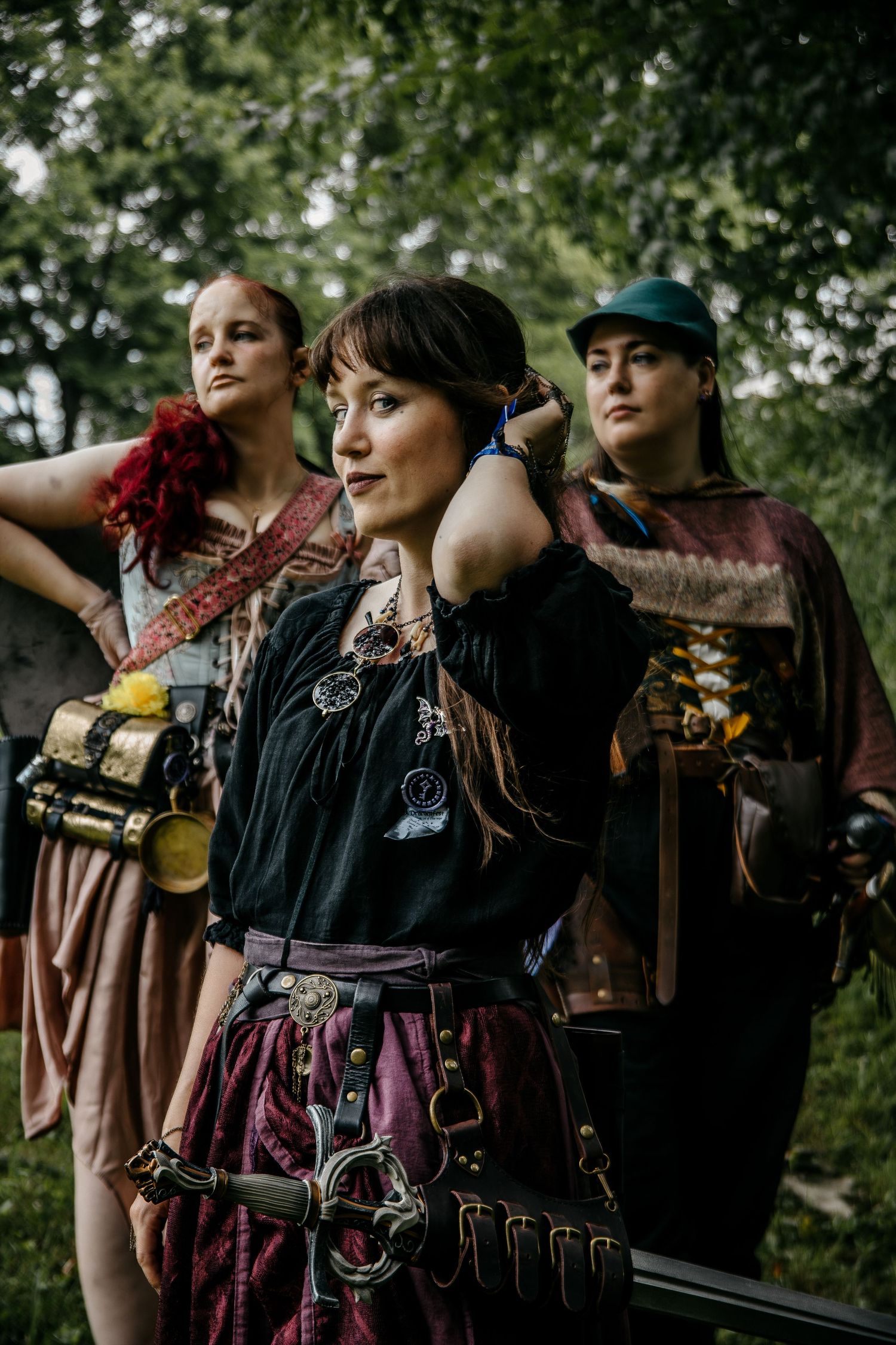 Three women in medieval fantasy costumes pose outdoors, wearing layered garments, leather accessories, and carrying props.