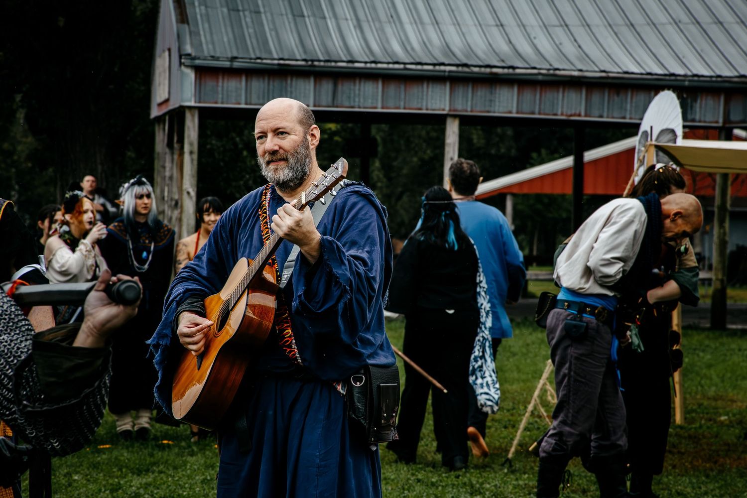 Man in medieval costume playing guitar at outdoor Renaissance faire with costumed participants in background.