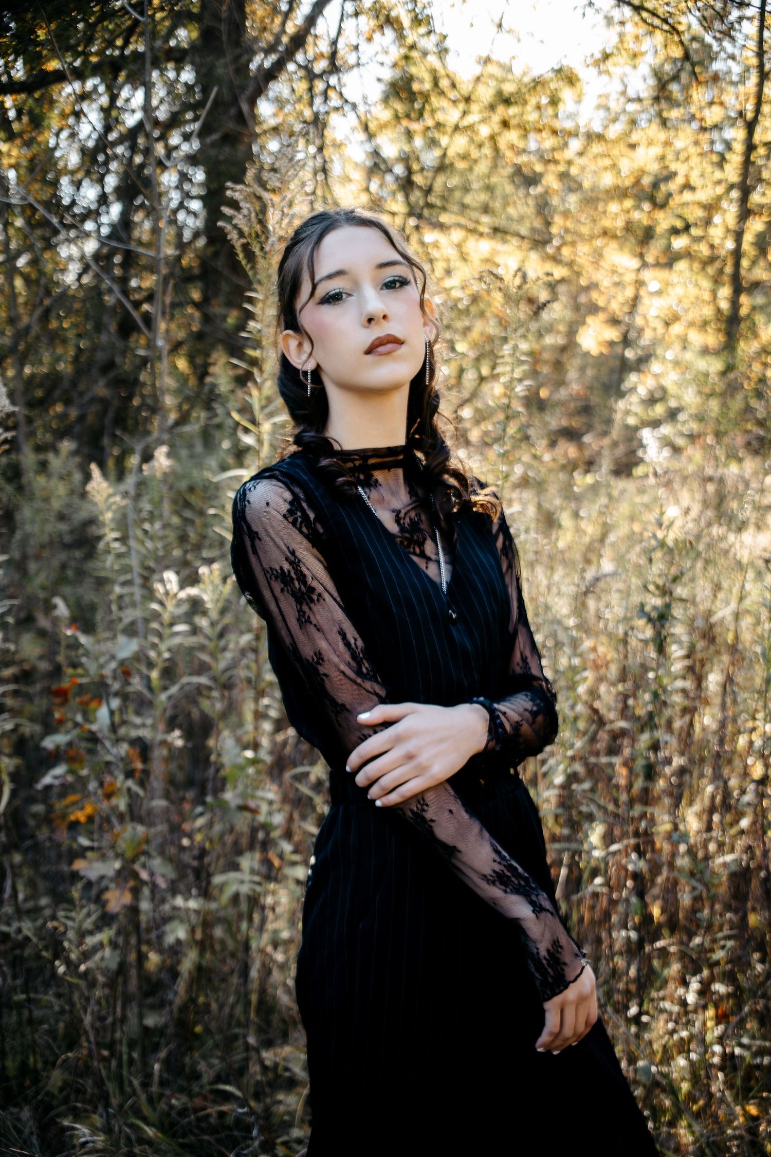 A woman in a black lace dress poses gracefully in a sunlit autumn forest setting.