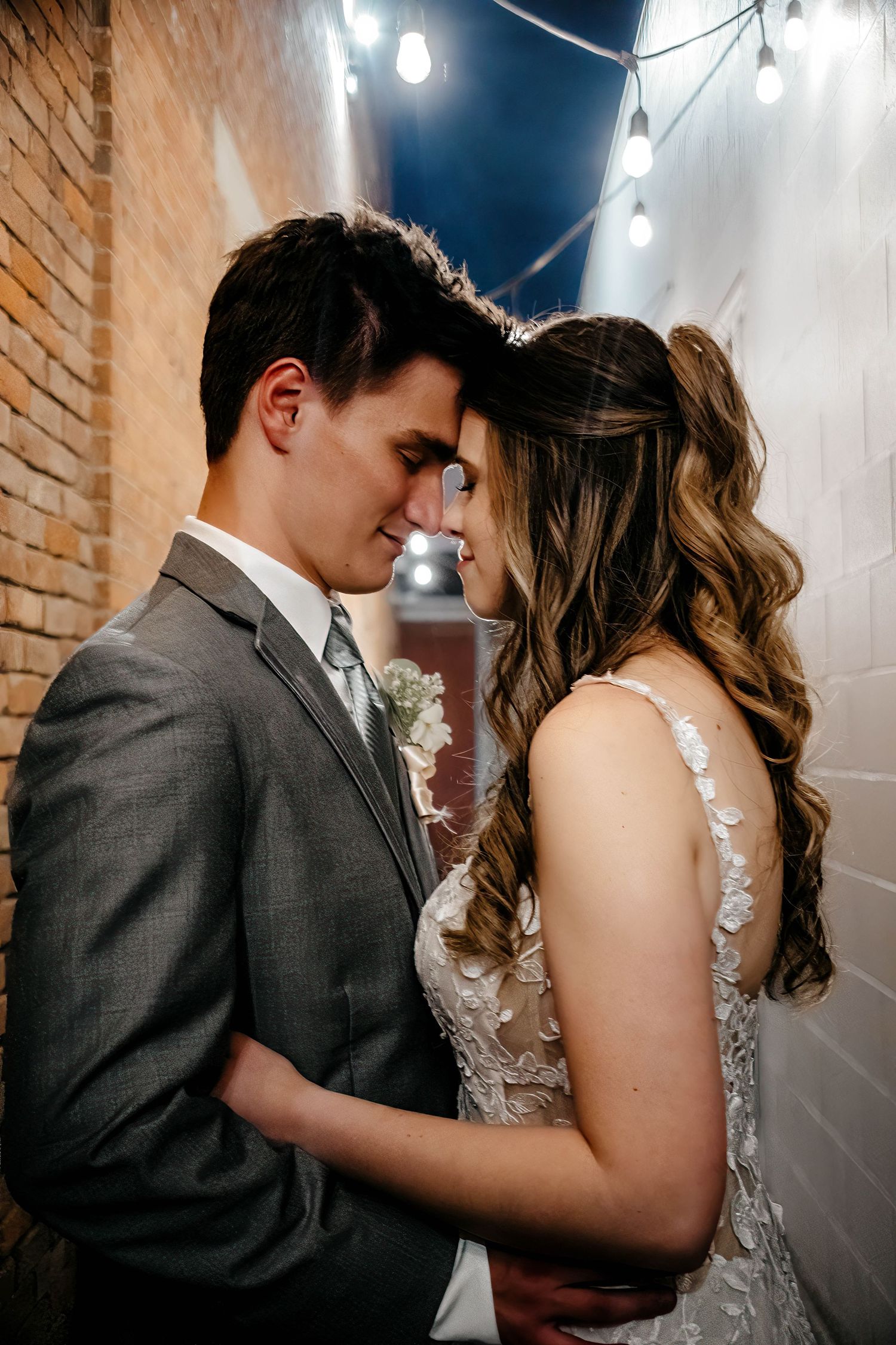 Romantic wedding couple sharing intimate moments in front of exposed brick wall under string lights.
