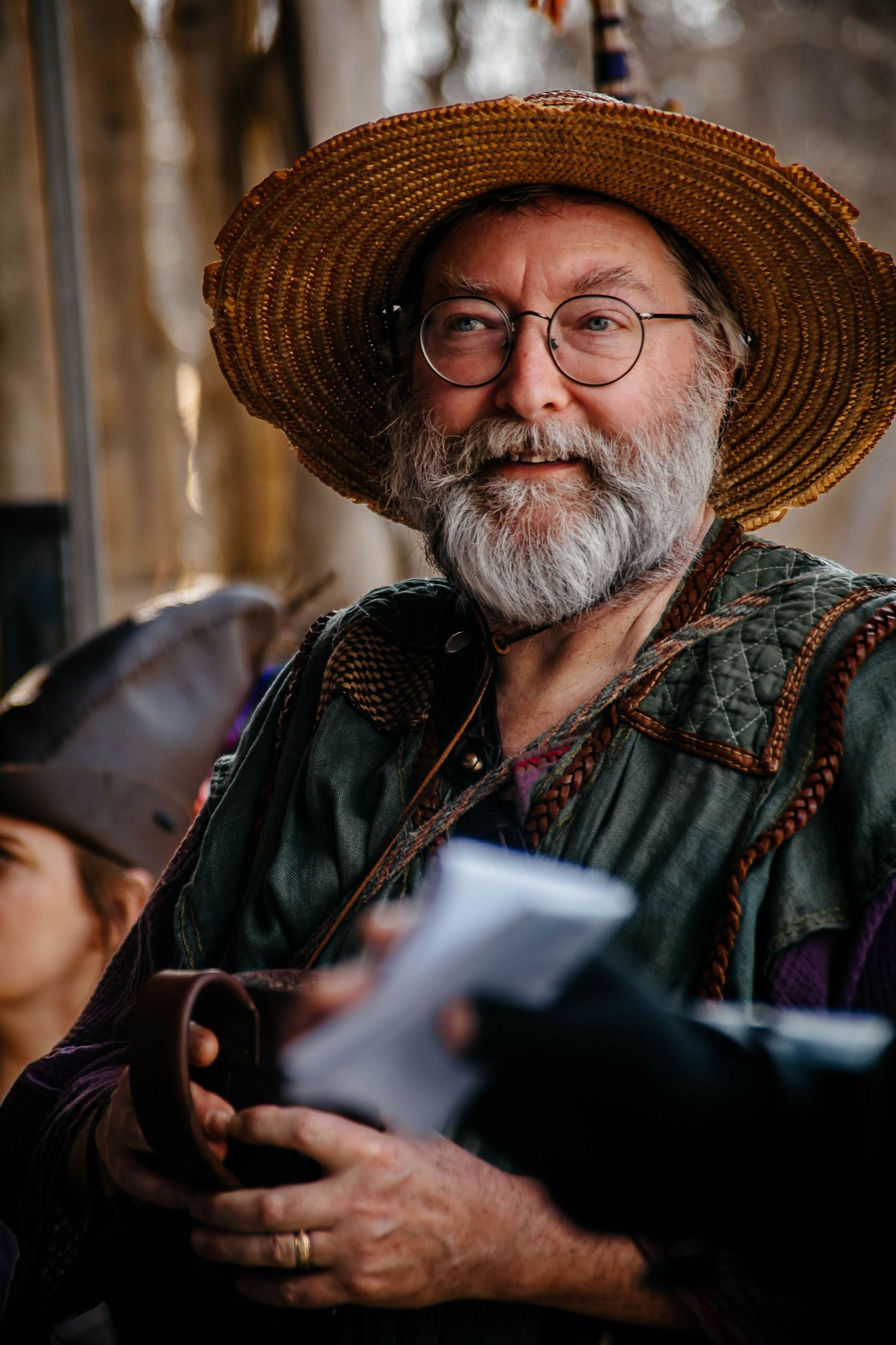 A bearded man in a straw hat and glasses holds a camera while smiling in a candid portrait series.