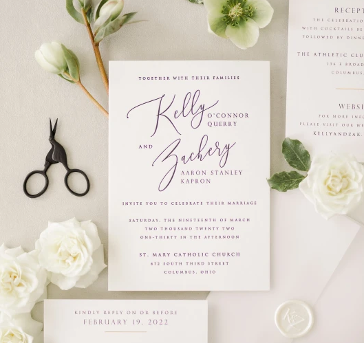 Wedding invitation suite with white roses, hellebores, and greenery arranged on a light beige background with scissors.
