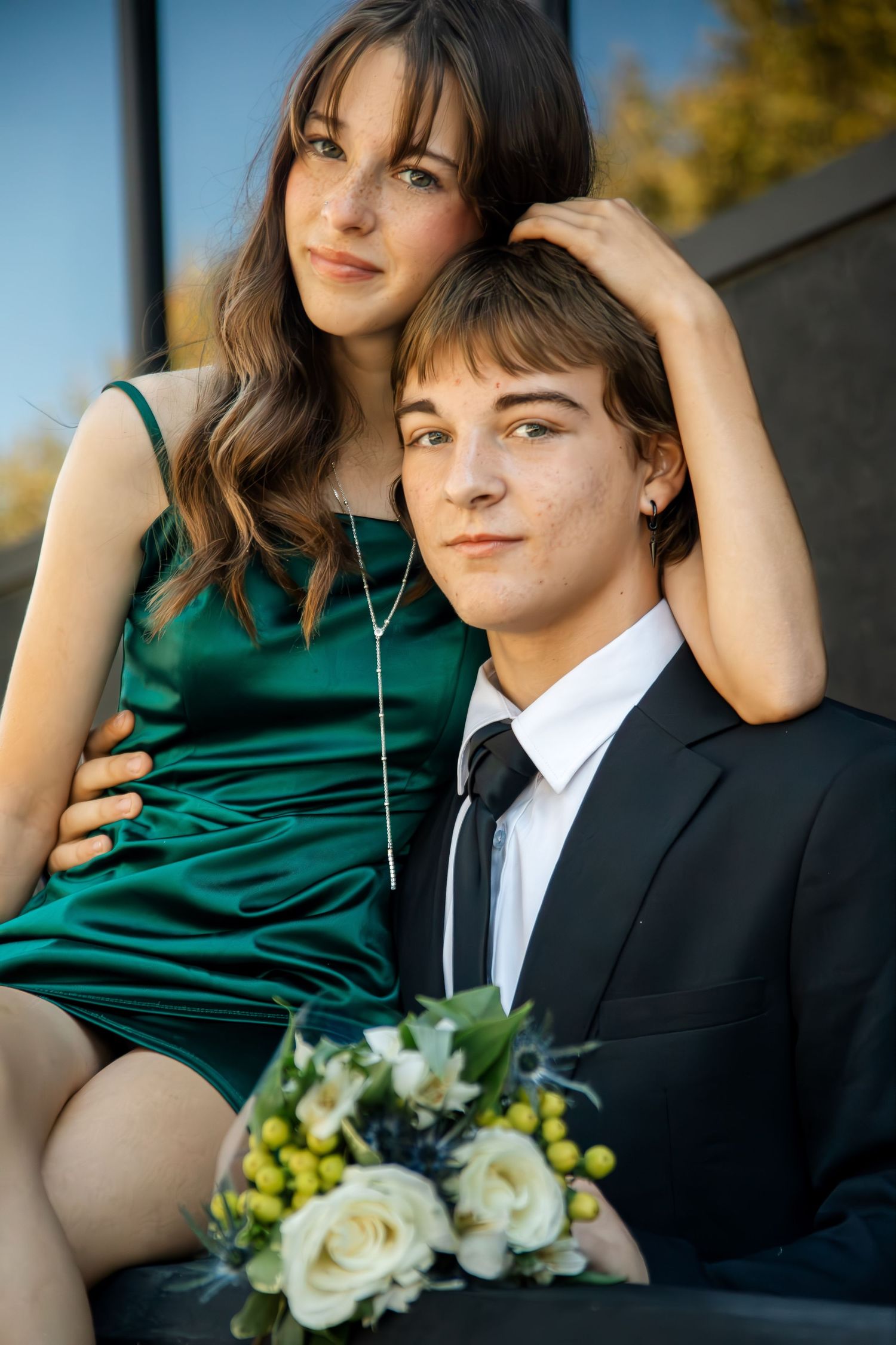 A couple in formal attire poses outdoors with a bouquet of white flowers against an autumn background.