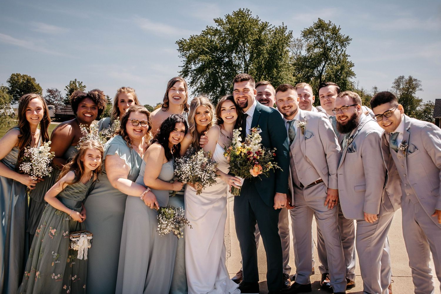 Large wedding party in neutral tones poses together outdoors under blue skies.