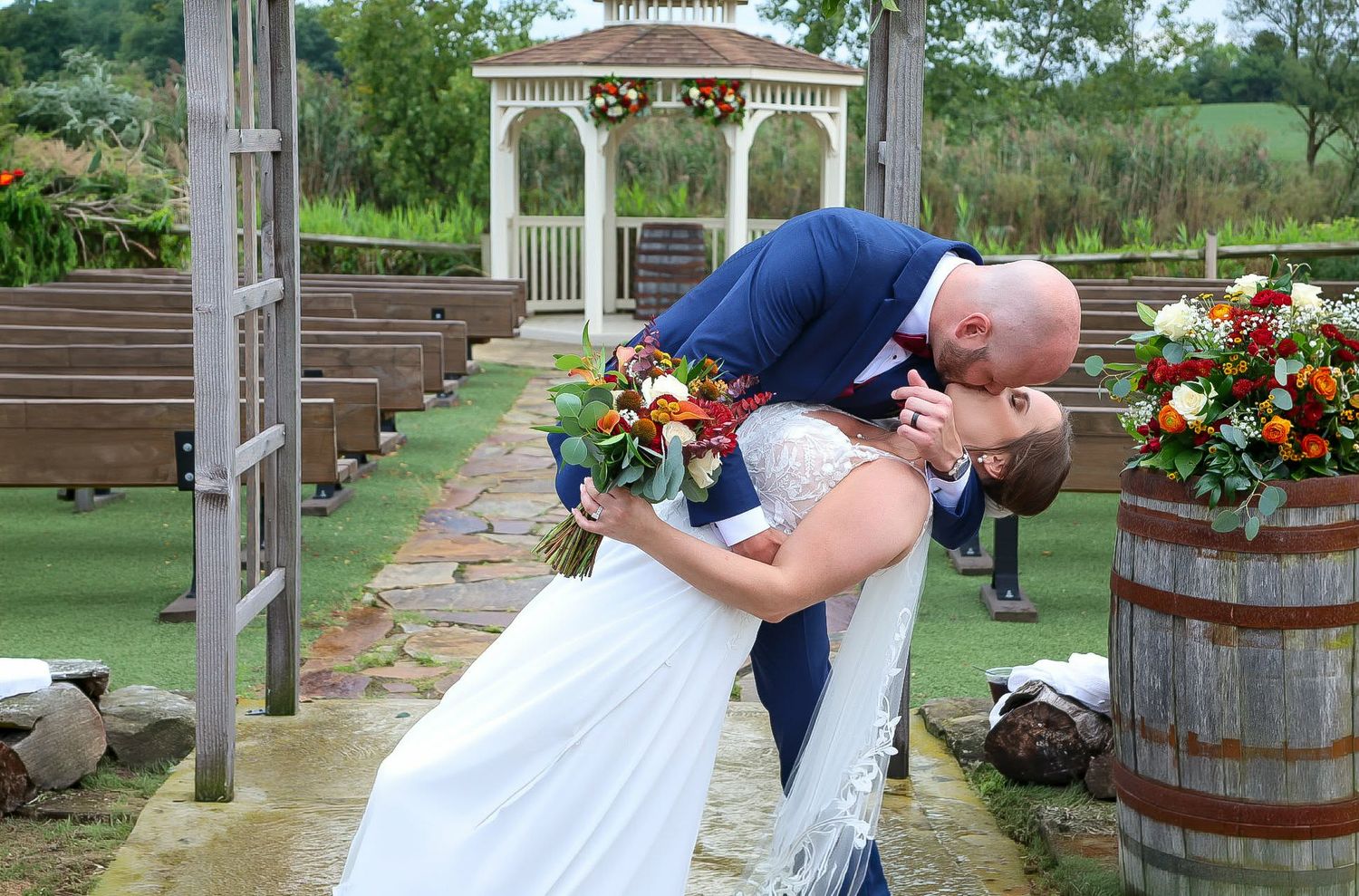A groom dips his bride for a romantic kiss at an outdoor wedding ceremony with a white gazebo backdrop.