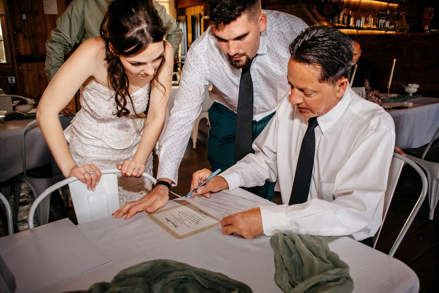 Wedding guests in white attire gather around tables to sign guestbooks and documents at a celebration.