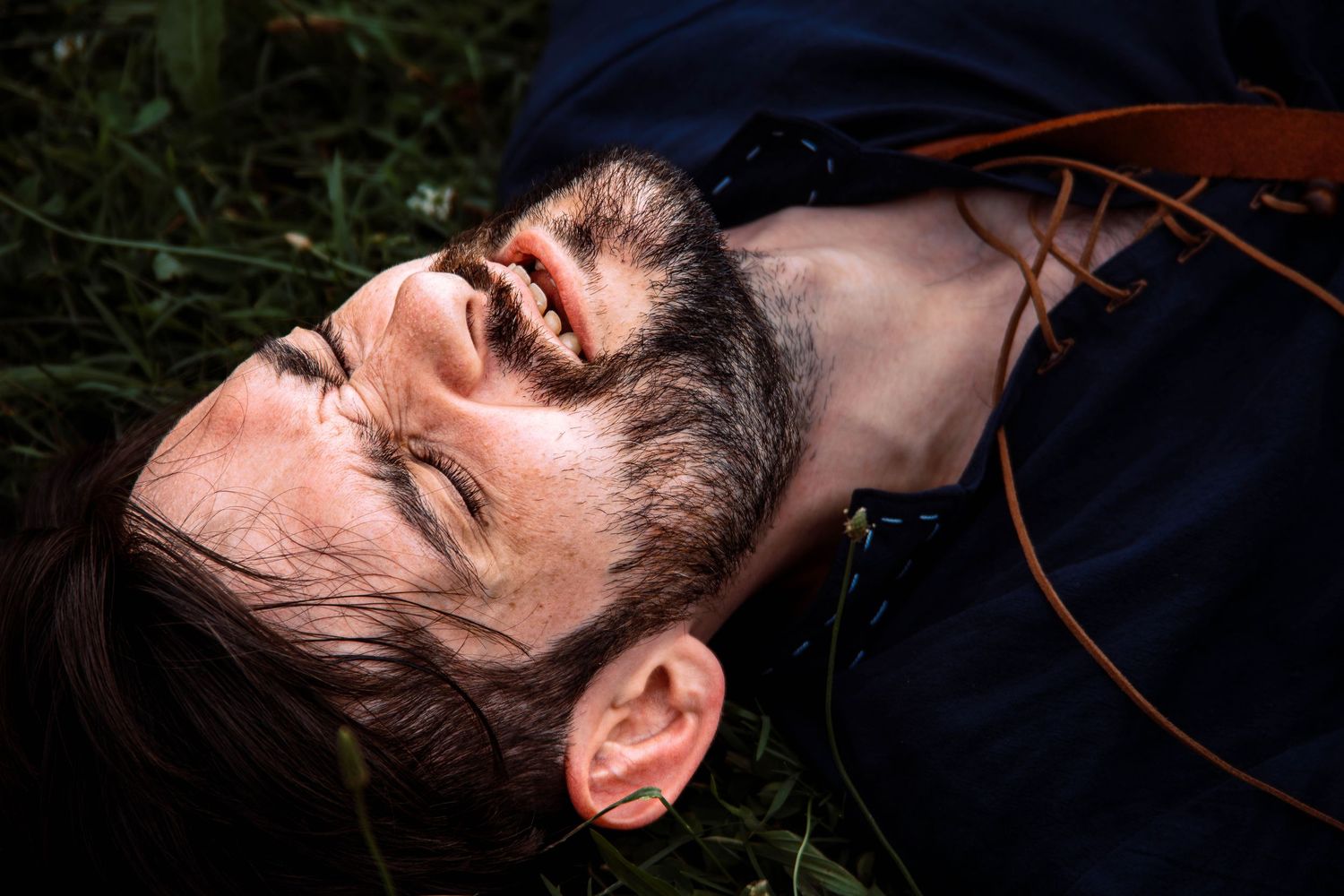 Person with dark hair and facial hair lies back with eyes closed wearing a dark colored shirt against a dark background.