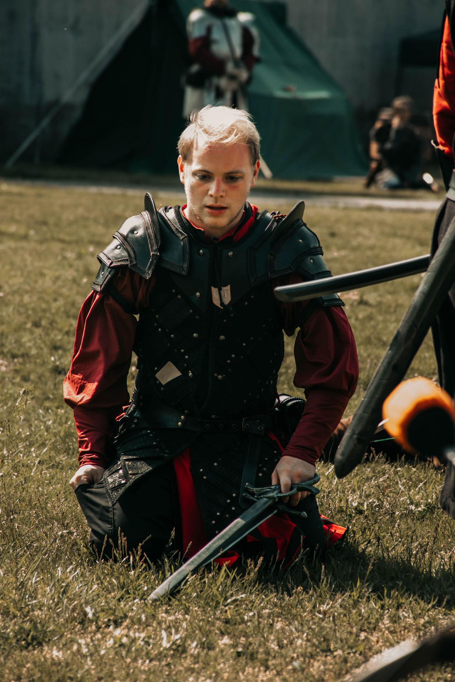 A person in medieval costume kneels on grass while holding a long sword during an outdoor reenactment event.