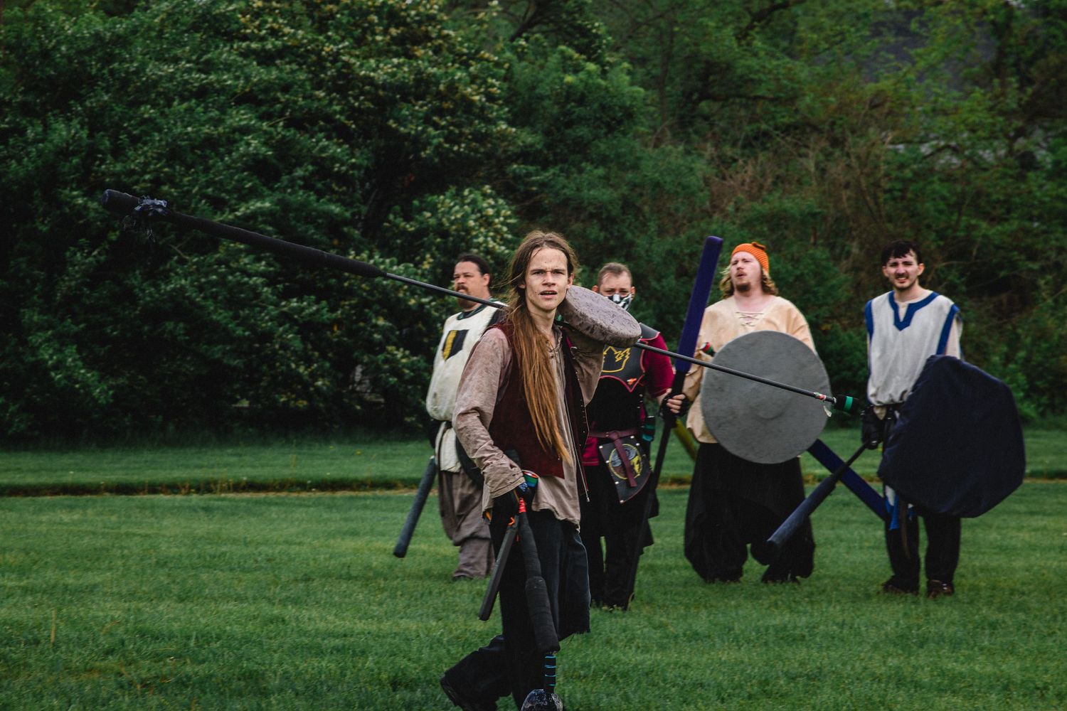 Medieval reenactors in costume battle with swords and shields on a green field surrounded by lush trees.