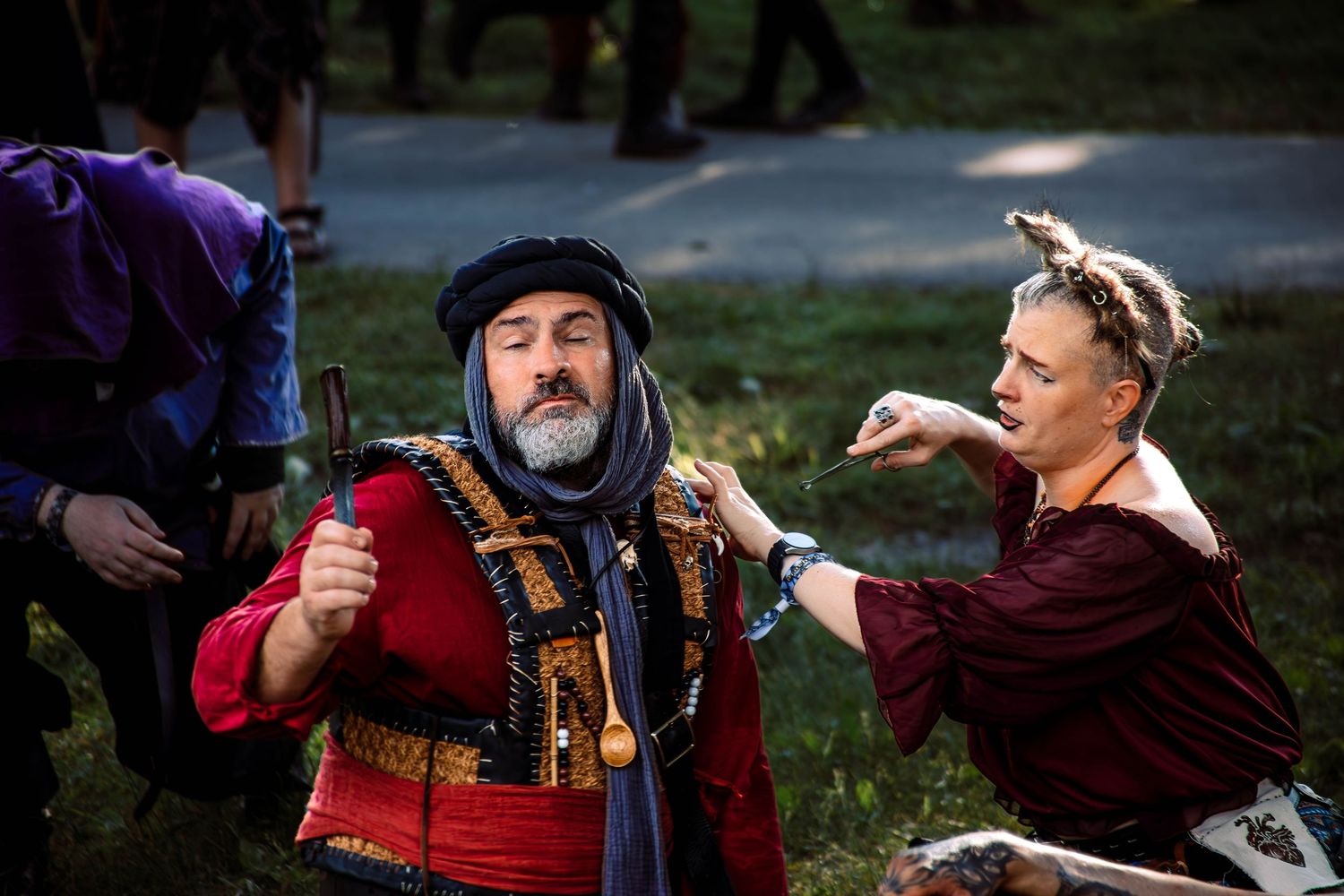 Two people in medieval Renaissance faire costumes, one in red and gold, interact outdoors at a historical reenactment event.