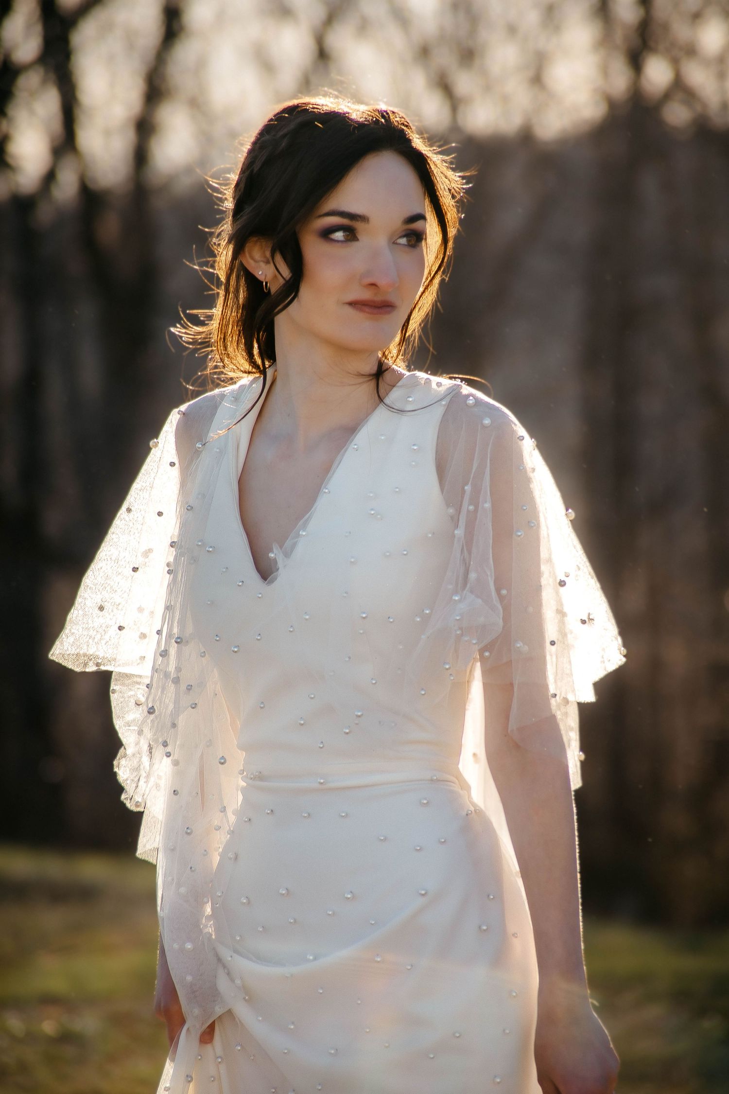 A woman in a romantic white dress with sheer flutter sleeves poses in golden sunlight against a wooded background.