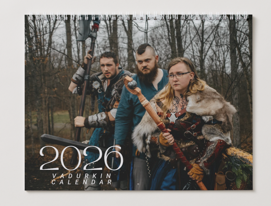 A group of people in winter clothing pose with medieval-style weapons in a wooded setting for a 2026 calendar photo.