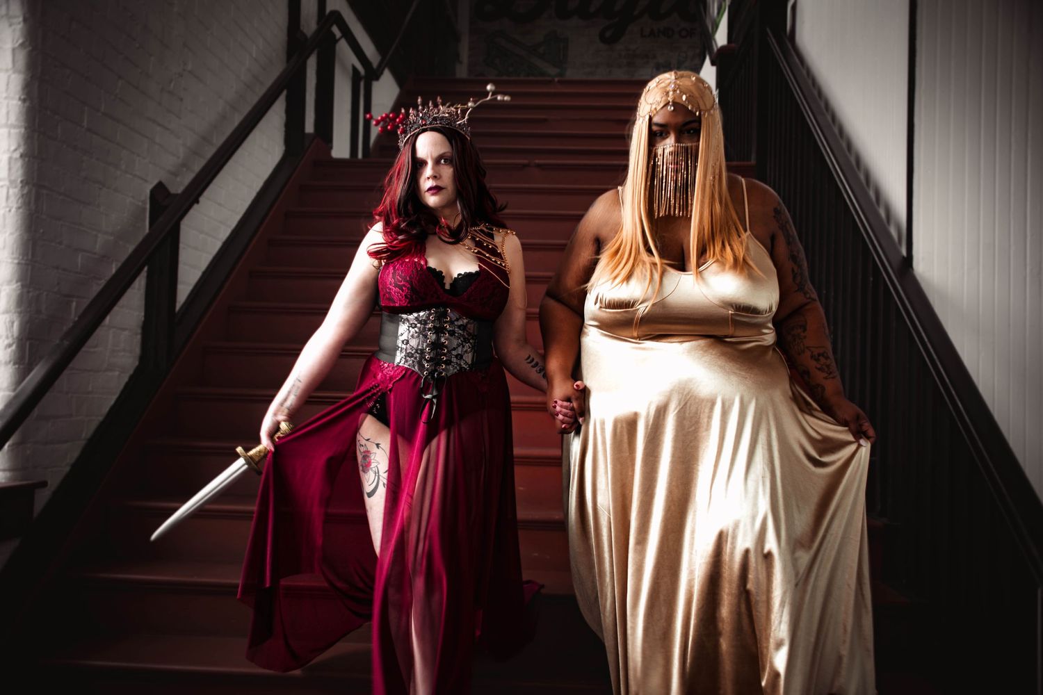 Two women in elaborate fantasy costumes pose on a staircase, one in red holding a sword, one in white.