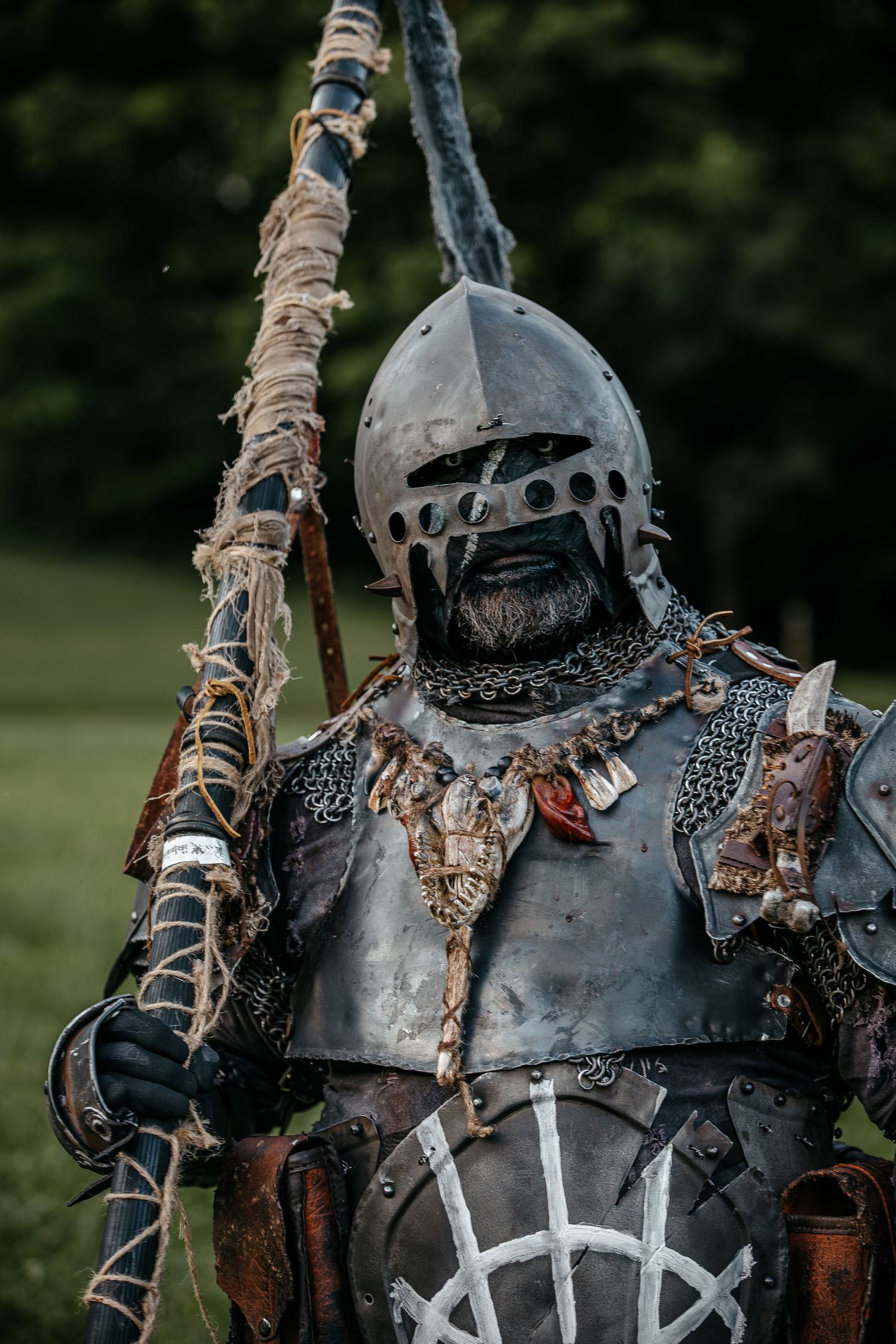 A warrior in dark medieval armor holds a weathered spear, wearing a metal helmet with chain mail outdoors.