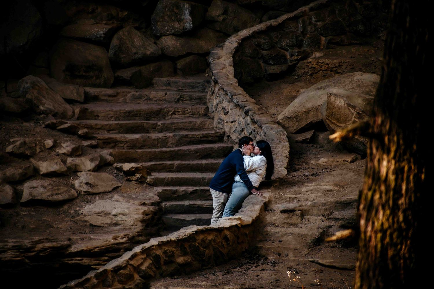 Couple sharing a romantic kiss on rustic stone steps surrounded by natural rock formations in an outdoor setting.