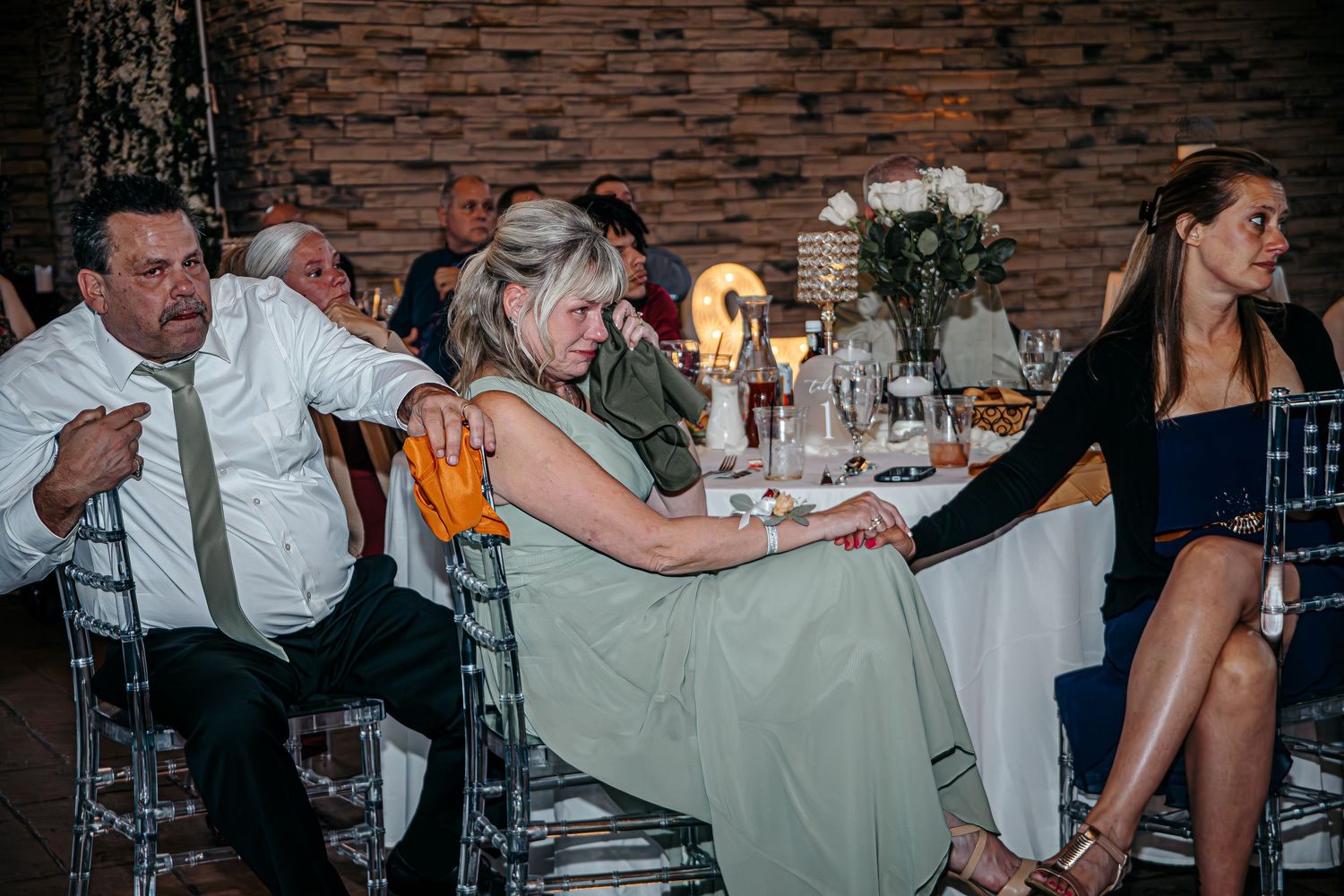 Guests seated at a wedding reception table in an elegant stone-walled venue with floral centerpieces.