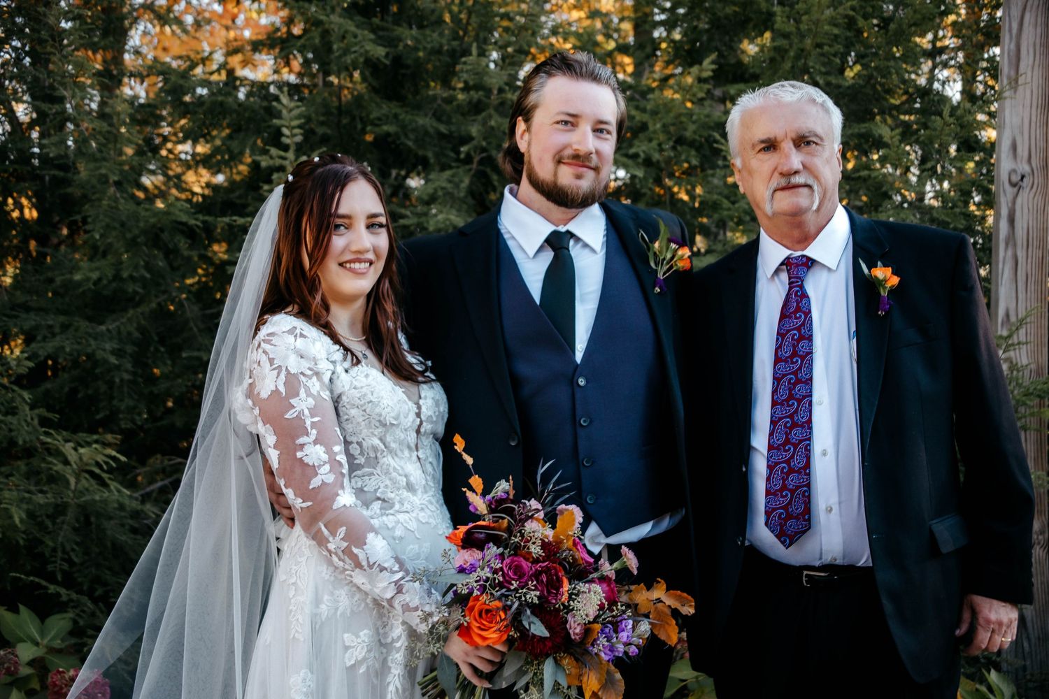 Wedding party poses together under autumn trees wearing formal attire and holding bouquets.