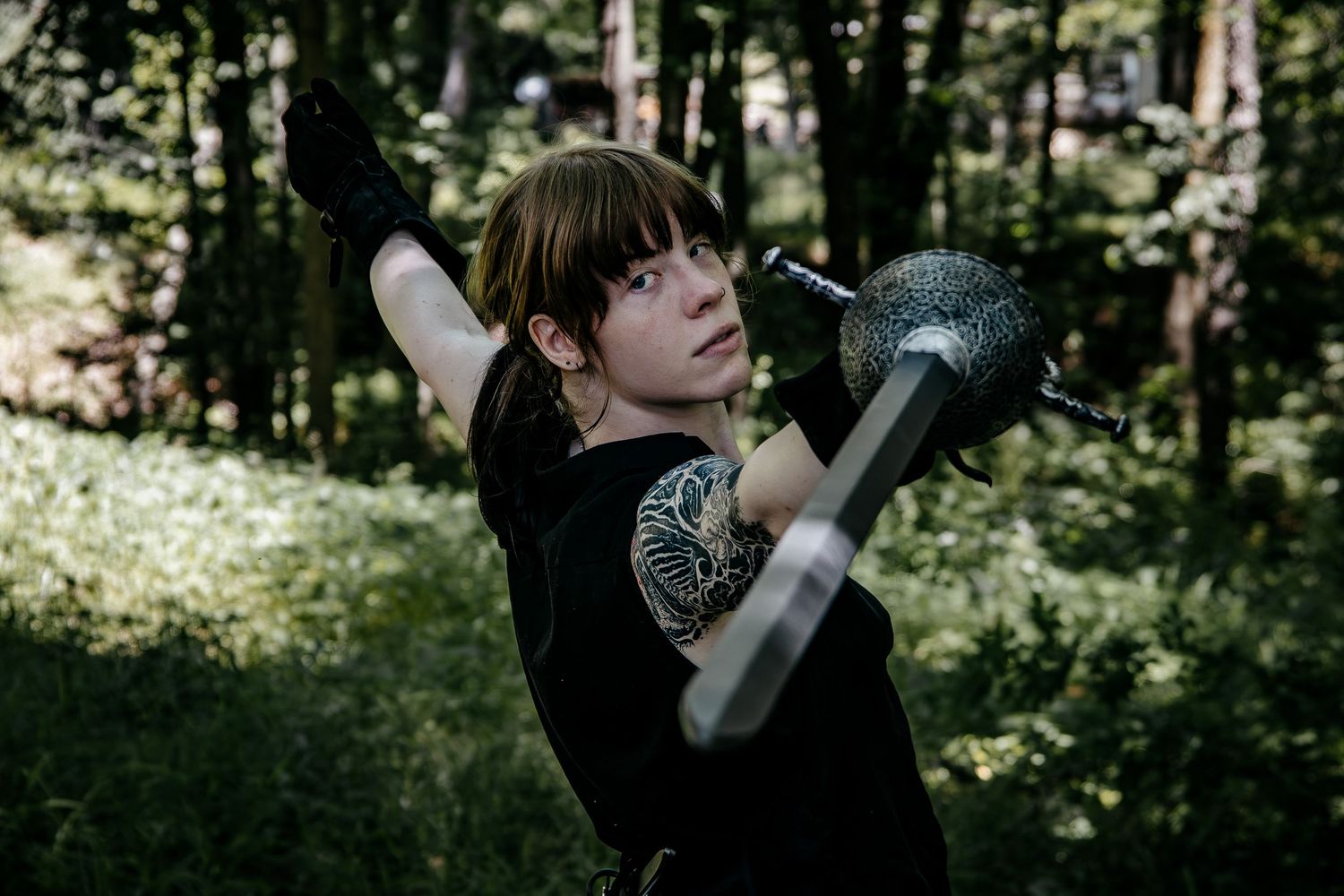 Young woman in black t-shirt poses with a decorative mace weapon outdoors in a lush green forest setting.