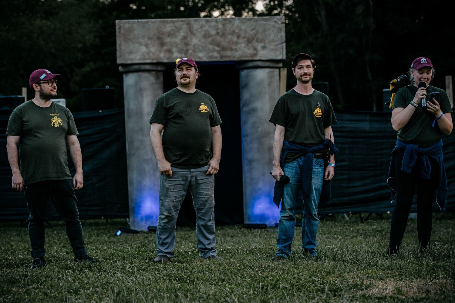 Four men in matching dark t-shirts standing outdoors near stone prop at evening festival event.