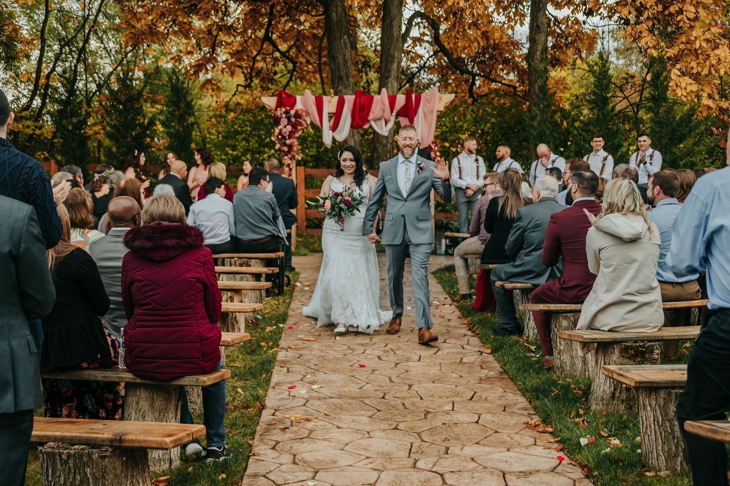 Outdoor autumn wedding ceremony with wooden benches and guests seated along a tree-lined path during golden hour.