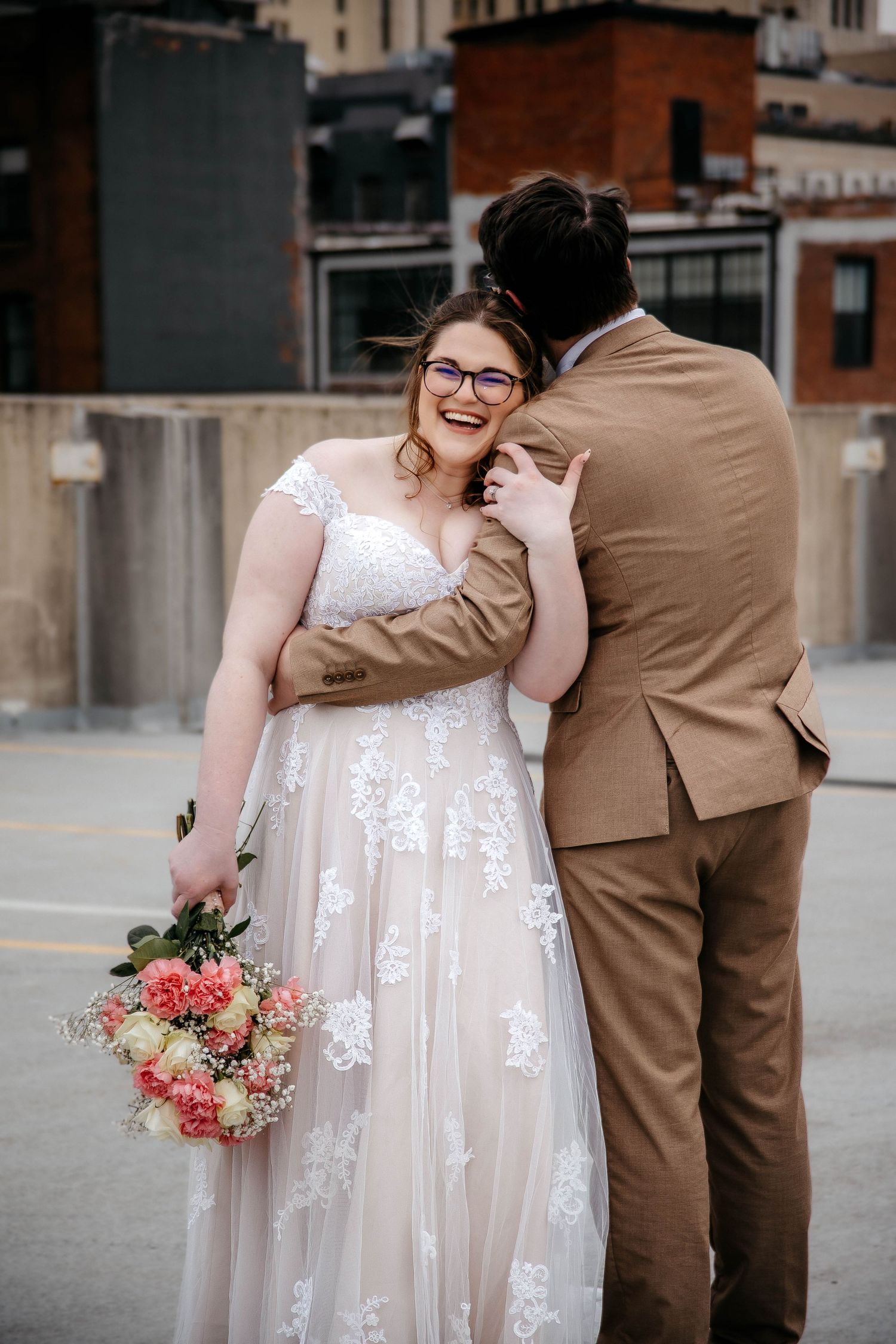 A romantic wedding moment captured in an urban setting with a brown suit and white lace dress against a city backdrop.