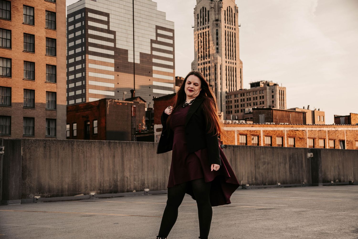 Person in black coat poses in front of urban buildings during golden hour sunset in city setting.