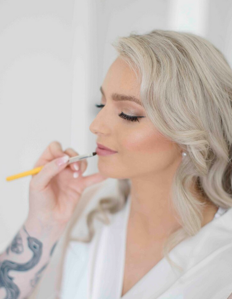 Makeup artist applies lipstick to a client wearing a white robe with a tattooed arm visible in the foreground.