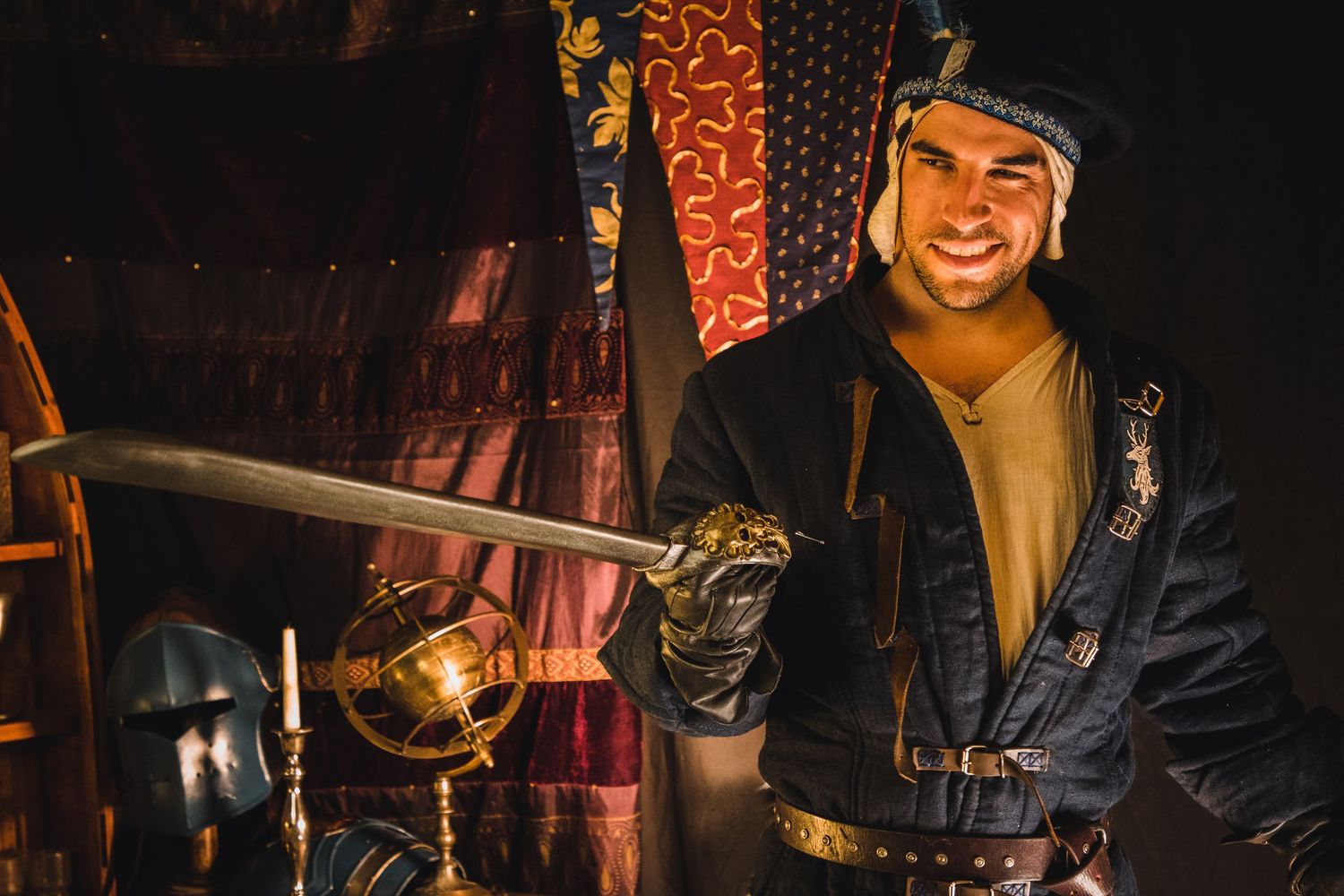 A performer in medieval costume smiles while holding a sword in a dimly lit theatrical setting.