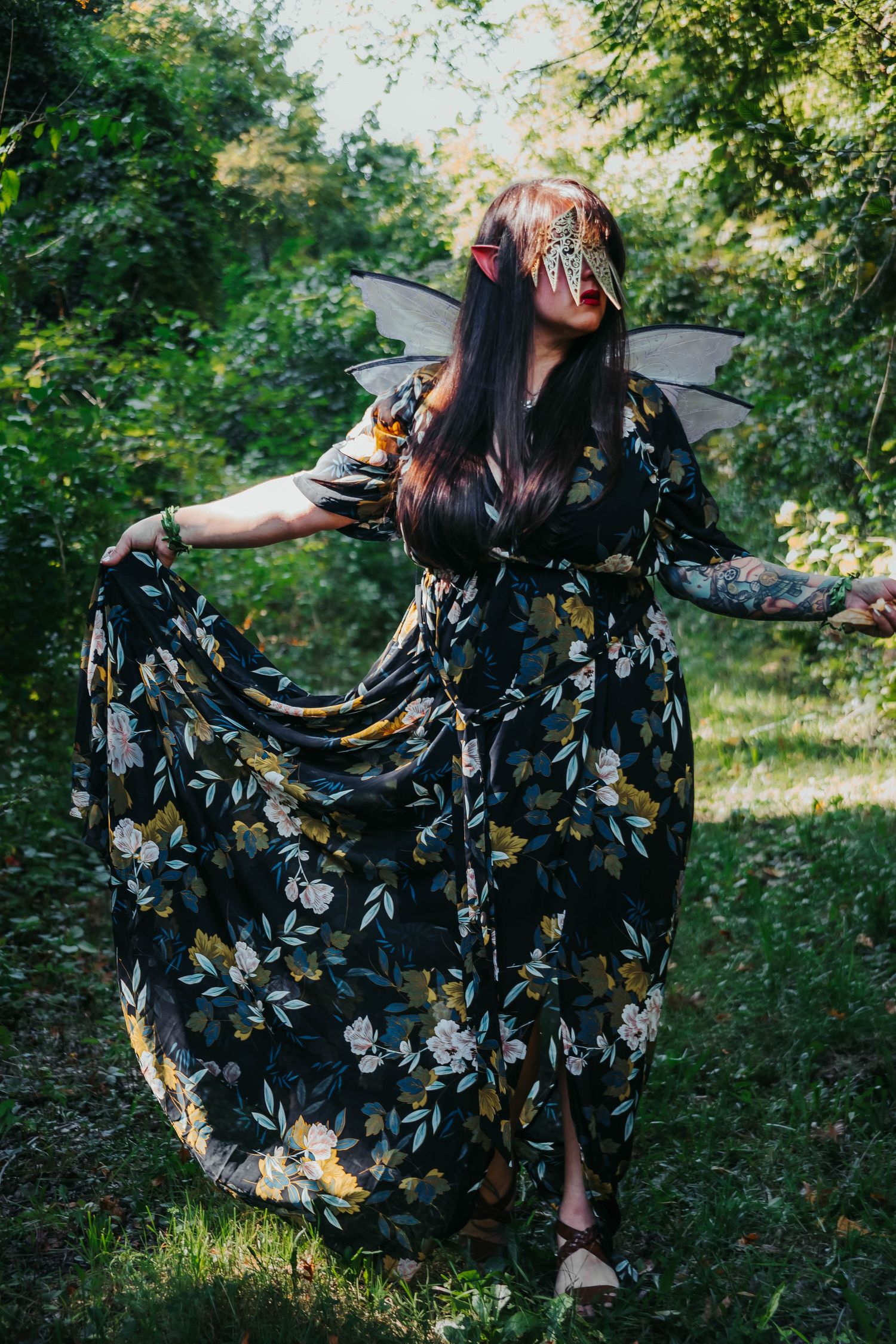 A figure in a flowing floral dress and fairy wings poses gracefully in a sunlit forest setting.
