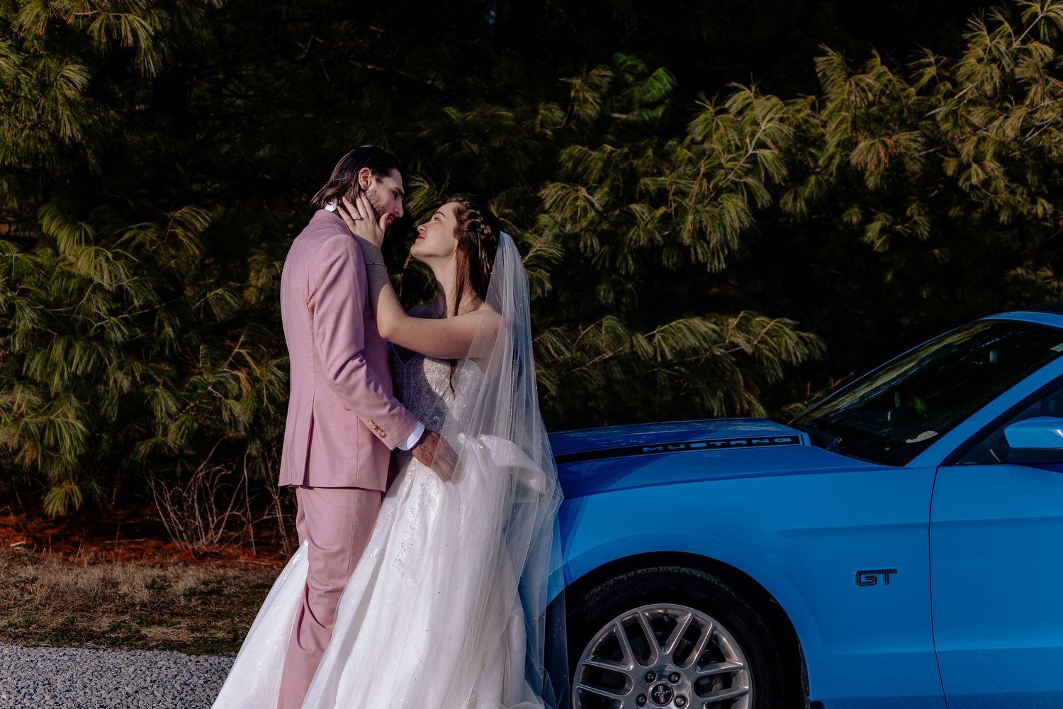 Bride and groom share a romantic kiss leaning against a blue Ford Mustang GT on their wedding day.