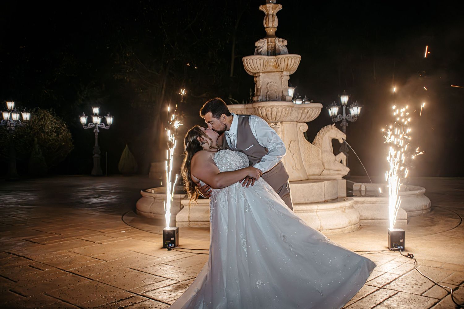 Bride and groom share romantic dip kiss near lit fountain with sparklers at night wedding celebration.