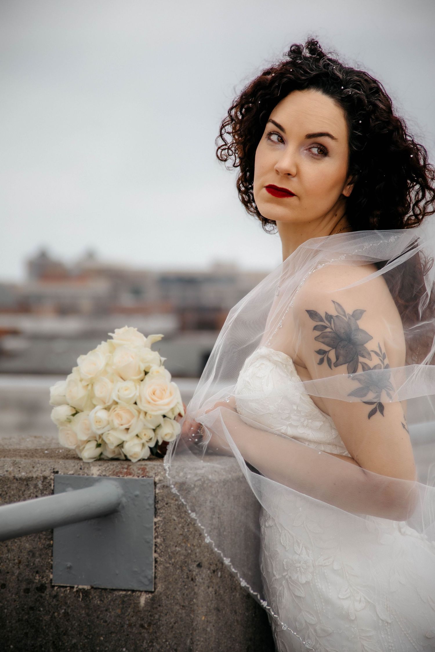 Bride in white lace gown and veil holds bouquet of white roses, showing floral tattoo on shoulder outdoors.