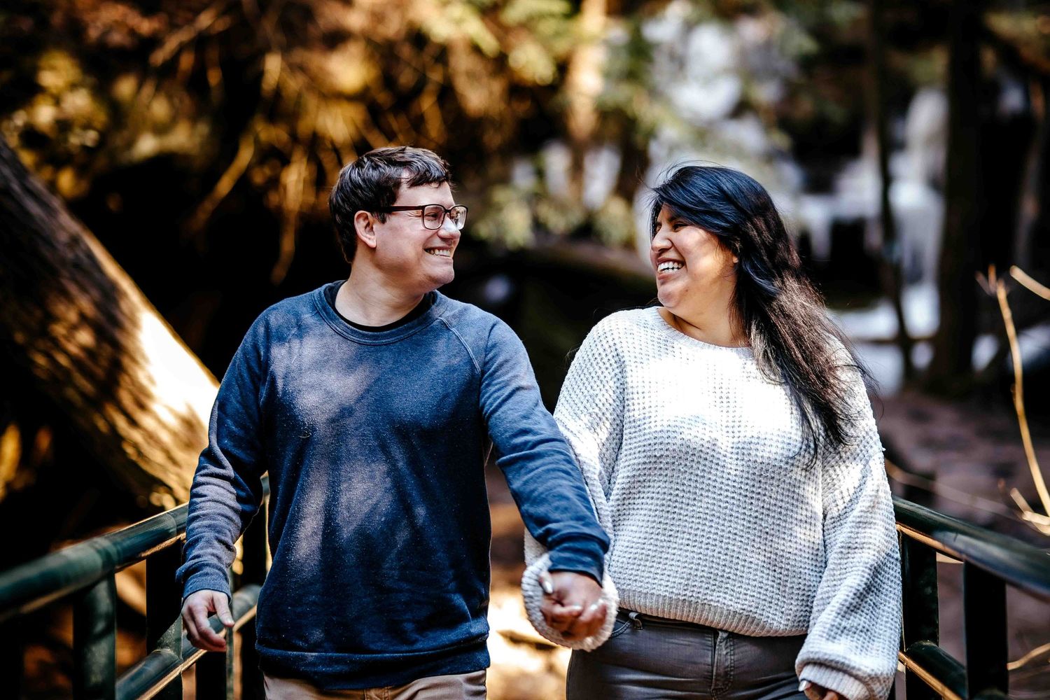 A happy couple holds hands and smiles at each other while walking on a bridge surrounded by trees.