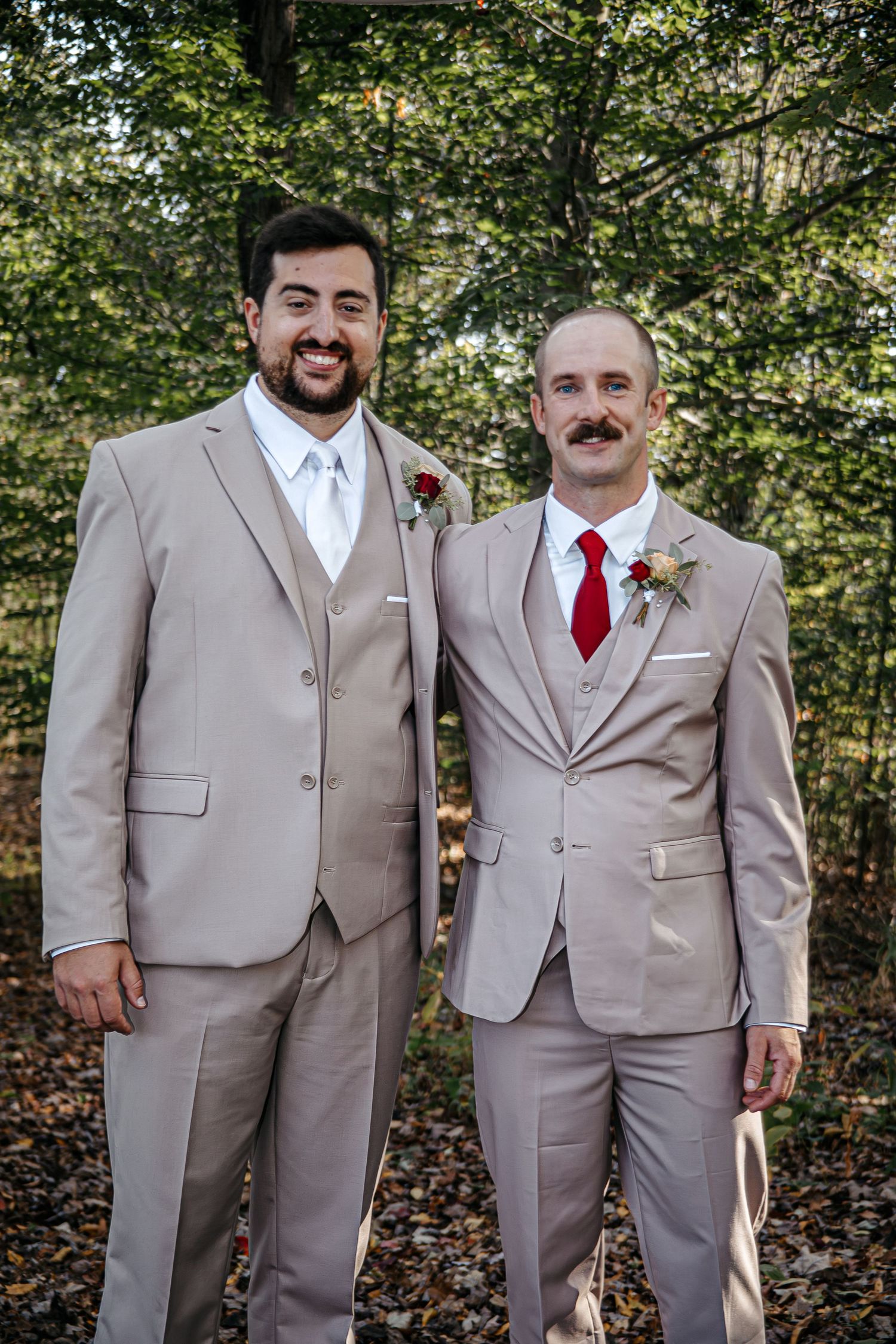 Groomsmen wearing matching tan suits and red ties pose together in a wooded outdoor setting.